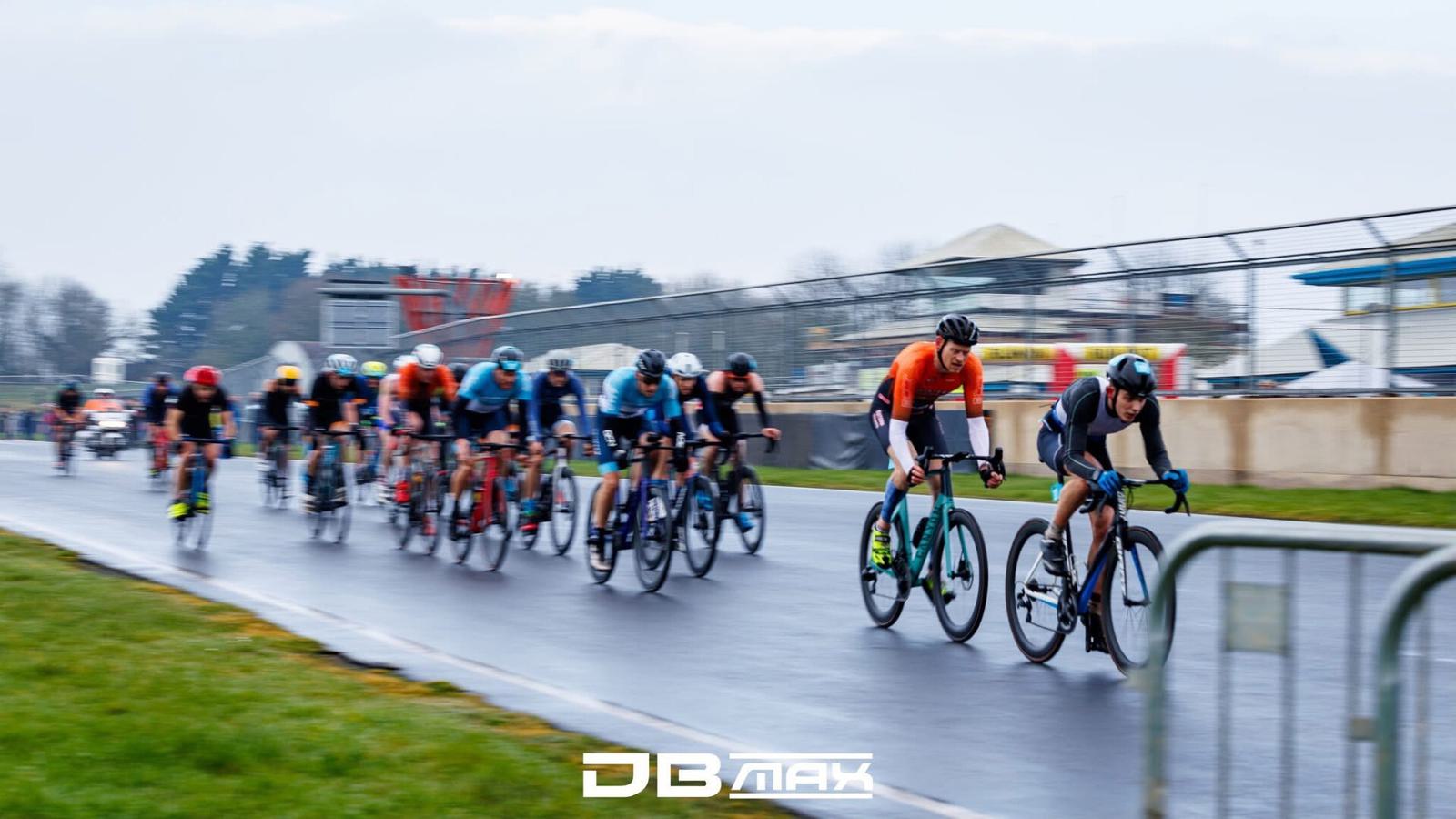 Cyclists compete in a road race on a wet track, wearing bright jerseys and helmets. Buildings and trees are visible in the background. The DB Max logo is at the bottom of the image.