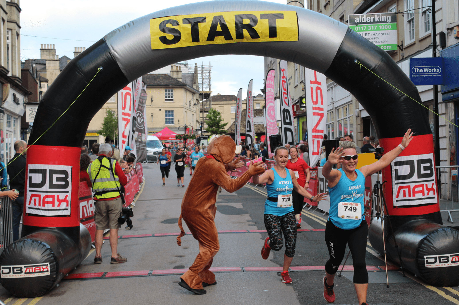Runners wearing race bibs, including one dressed in a bear costume, celebrate as they cross the start line of a race. A large black and yellow "START" arch marks the beginning of the race. Spectators and an official in a reflective vest are visible in the background.