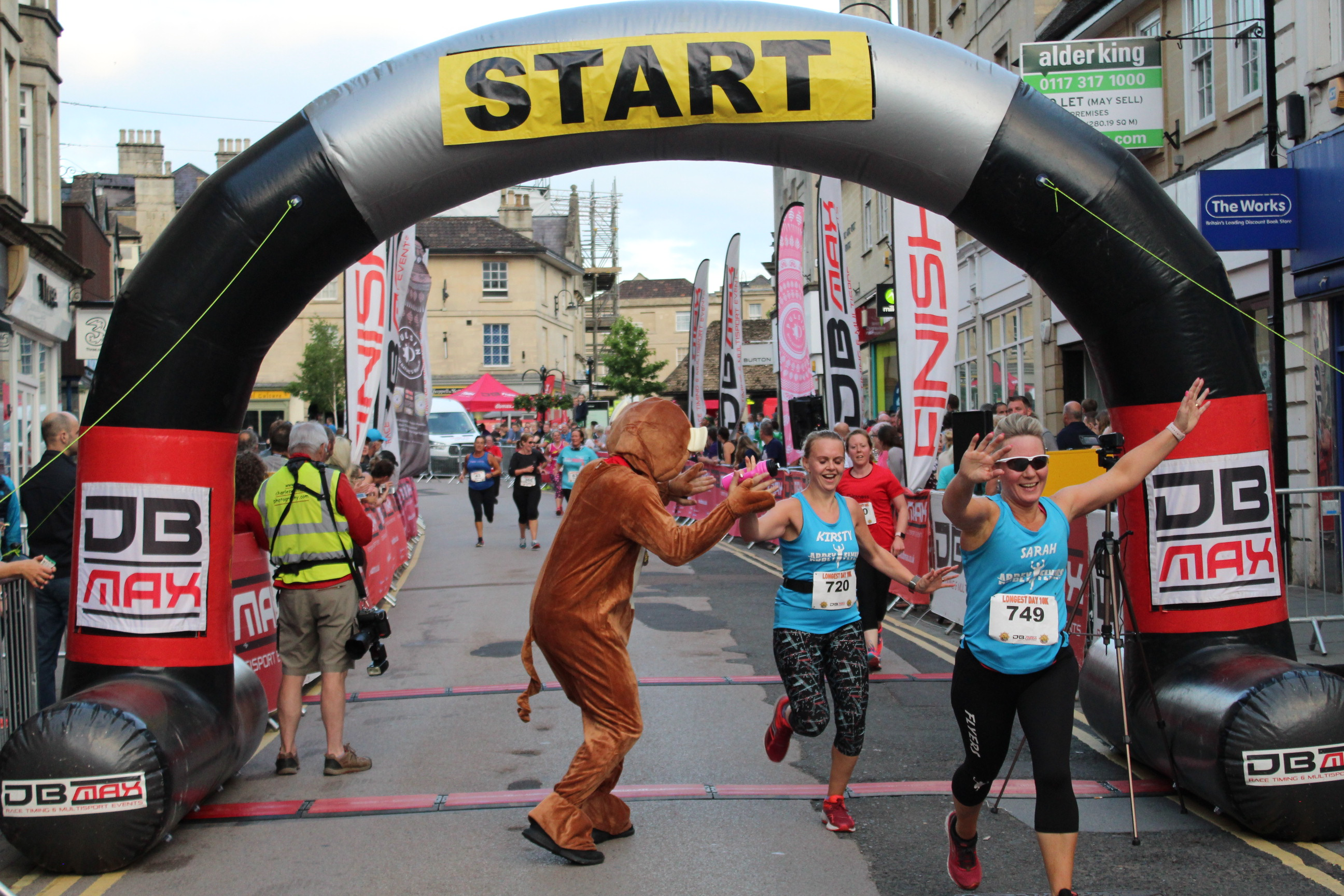 Runners wearing race bibs, including one dressed in a bear costume, celebrate as they cross the start line of a race. A large black and yellow "START" arch marks the beginning of the race. Spectators and an official in a reflective vest are visible in the background.