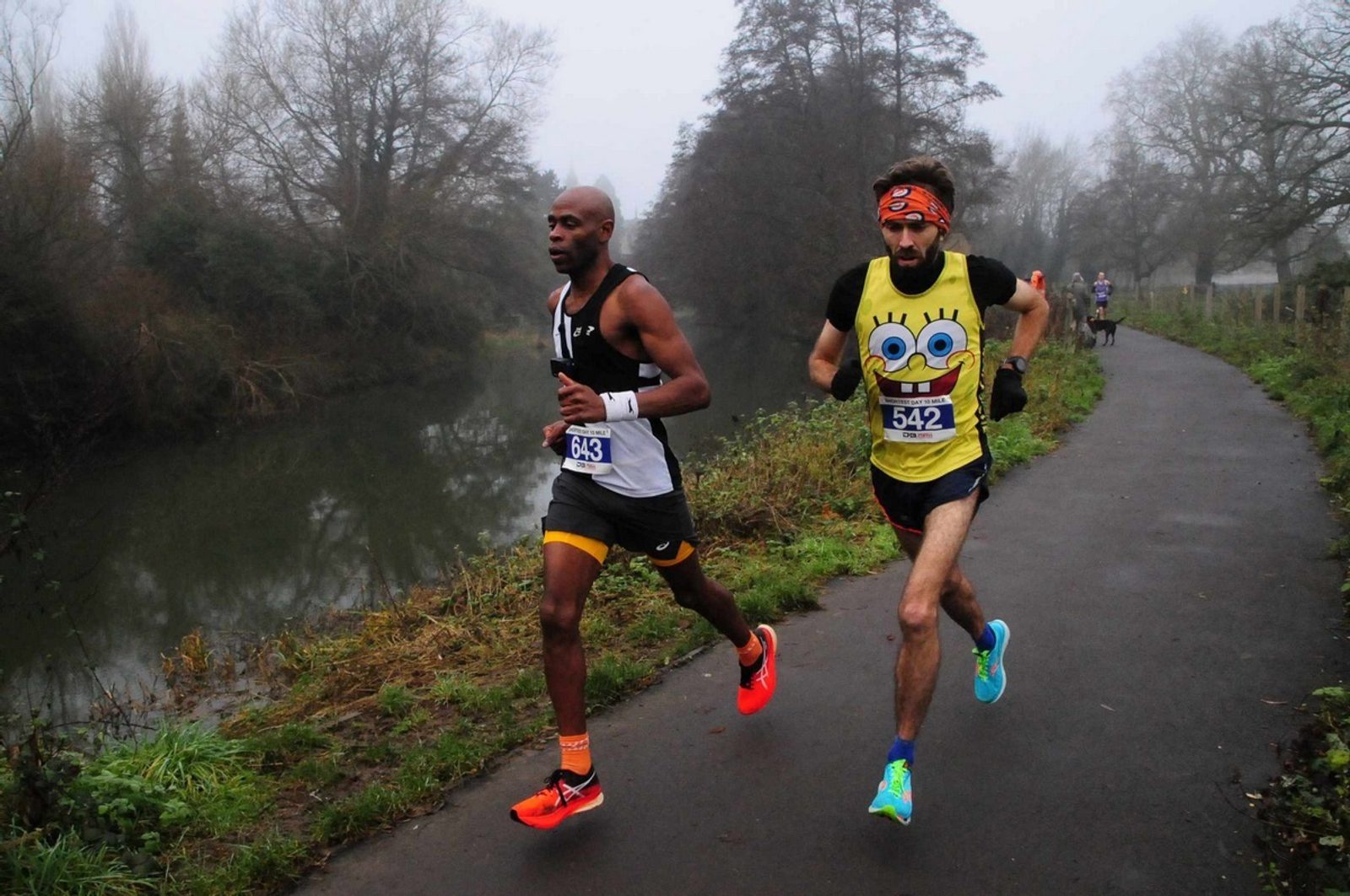 Two runners are participating in a race on a path next to a river on a foggy day. One runner wears a black outfit with orange shoes, and the other wears a yellow SpongeBob SquarePants shirt and bright blue shorts. The path is lined with trees and grass.