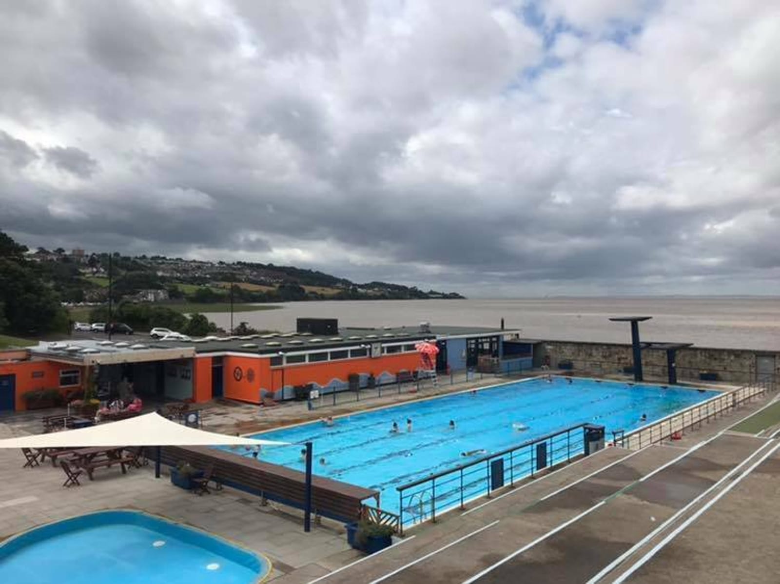 A coastal outdoor swimming pool with a few people swimming and relaxing. The pool area has colorful buildings and seating areas. Beyond the pool, there's a view of the ocean and hilly landscape under a cloudy sky.