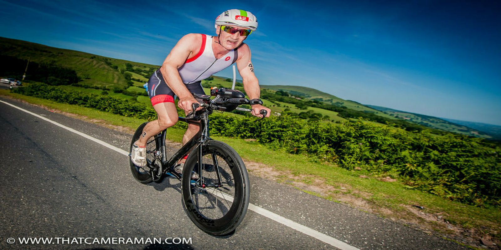 A cyclist wearing a white and red sleeveless cycling outfit and helmet rides a black bicycle on a road flanked by lush green hills under a clear blue sky. The image includes the watermark "WWW.THATCAMERAMAN.COM" at the bottom.