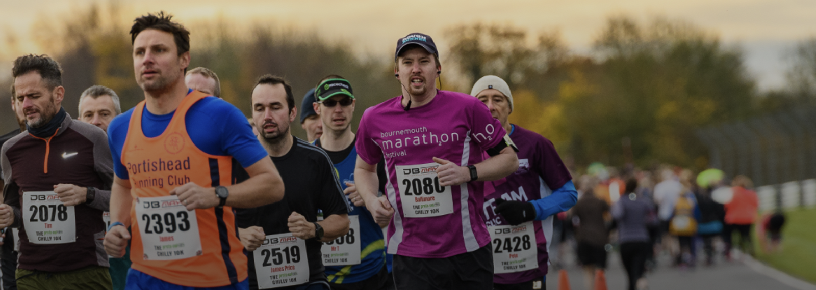 A group of runners participating in a marathon. One runner in an orange shirt with the text "Portishead Running Club" stands out among the others. The race is happening on a city street with trees and a sunset in the background.