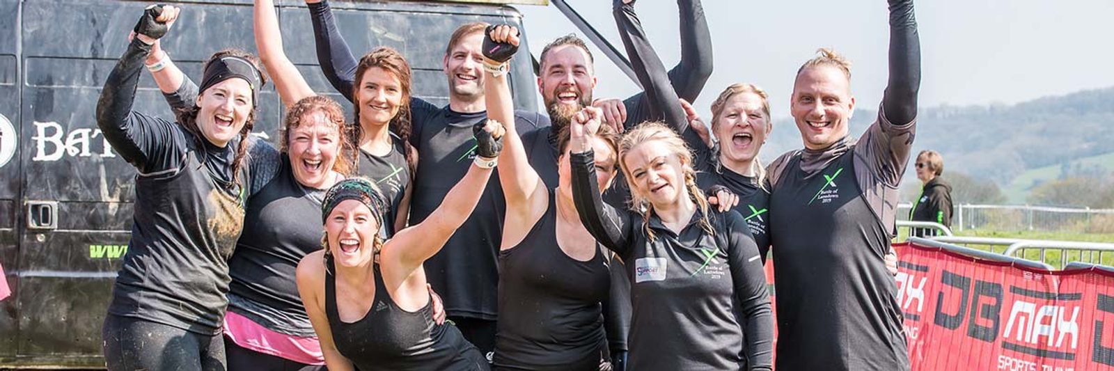 A group of people wearing black athletic gear smiling and cheering with their hands raised. They appear to have just completed a challenging outdoor activity, standing next to a large vehicle and a red barrier. The background features a hilly landscape.