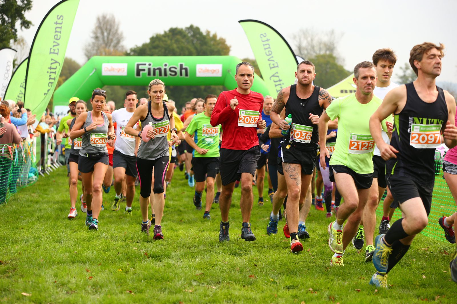 A group of runners approaching a green finish line arch at a charity event. They are wearing race bibs with numbers and are running on a grassy field. Spectators stand behind a fence on the left, and trees are visible in the background.