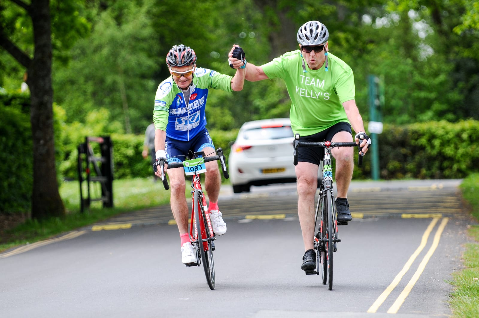Two cyclists participating in a race share a high-five while riding. The cyclist on the left is dressed in a blue and white outfit and the cyclist on the right wears a green shirt with "TEAM KELLY'S" on it. They are on a paved road surrounded by lush greenery.