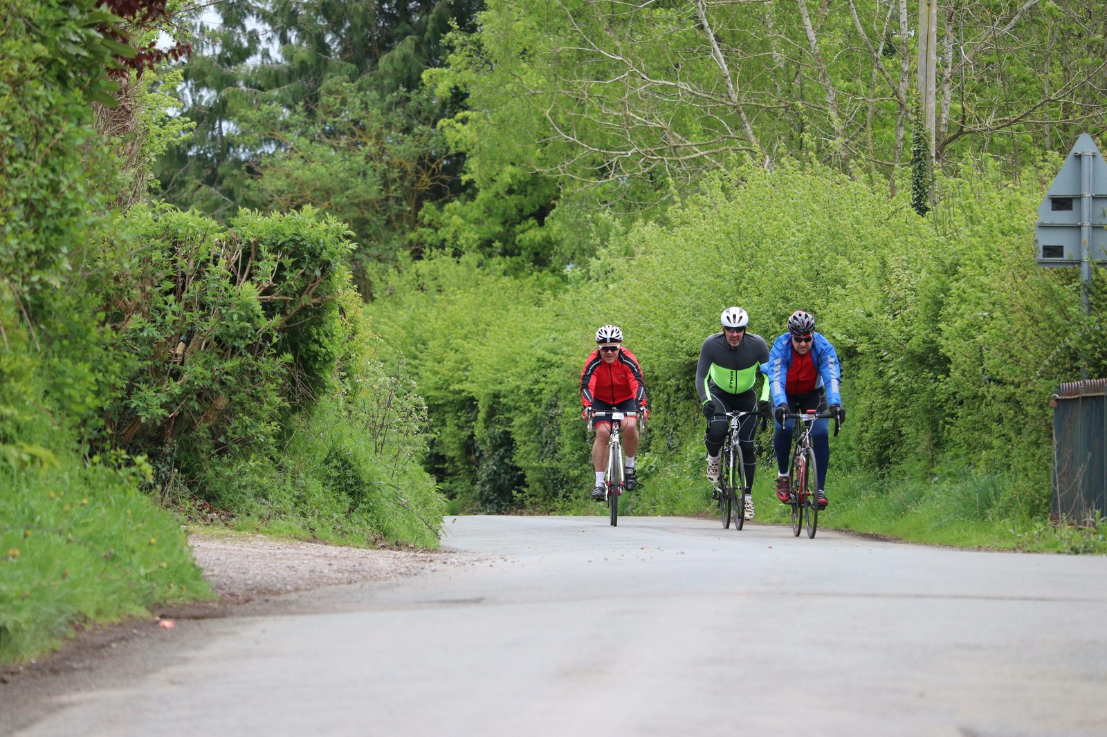 Three cyclists wearing helmets and colorful cycling gear ride together on a quiet road surrounded by lush greenery. The road curves slightly to the left, and the cyclists appear focused on their ride. A signpost is partially visible on the right side of the image.