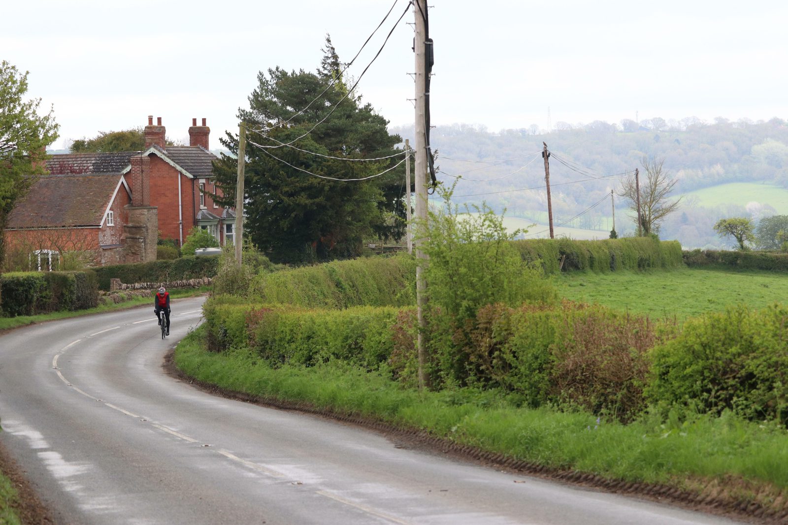 A cyclist rides along a curving rural road bordered by lush green hedges and fields. A few red brick houses and large trees are visible on the left side of the road, with rolling hills in the distant background under a cloudy sky.