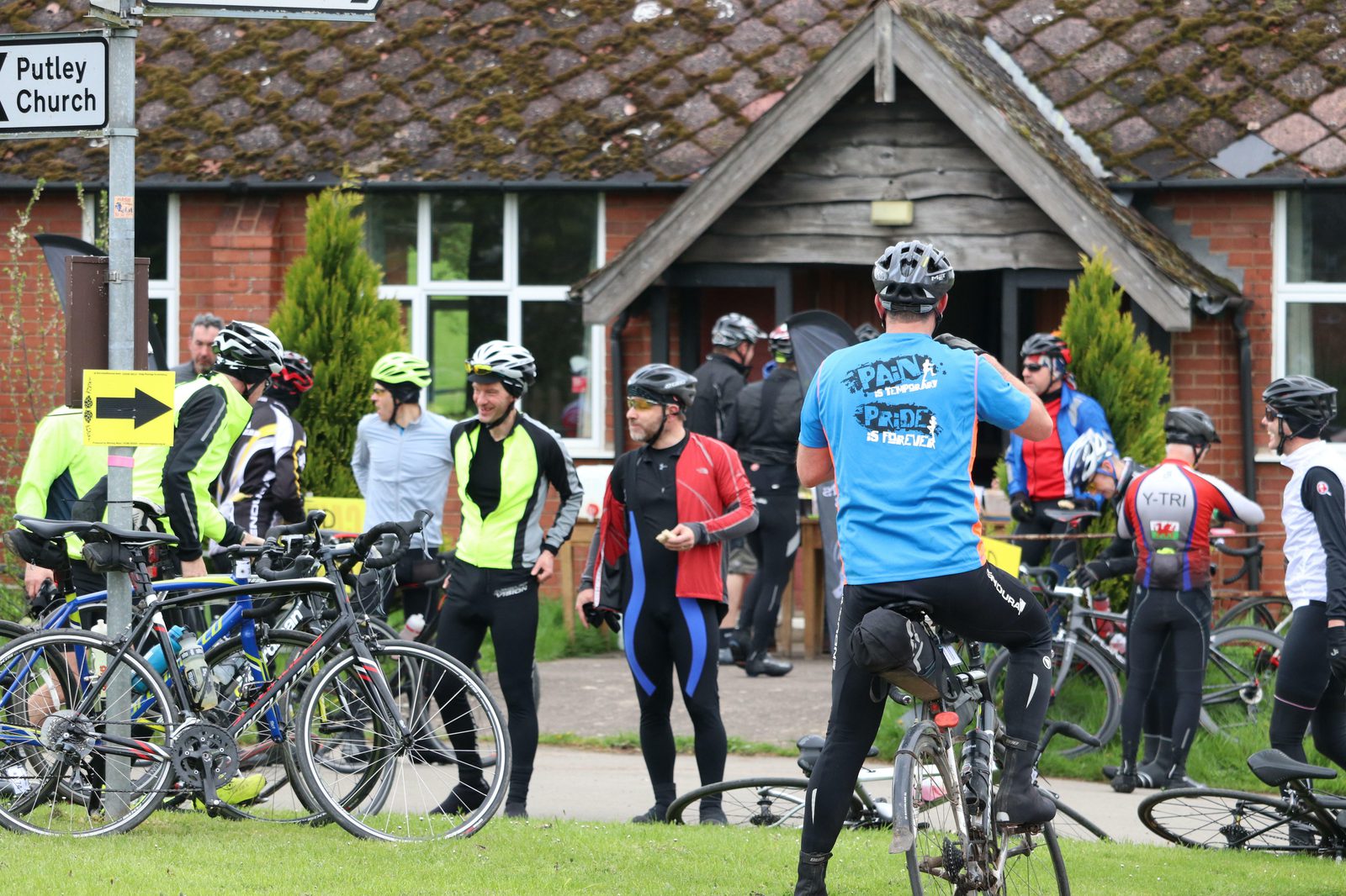 A group of cyclists wearing various colorful gear gather outside a rustic brick building with a wooden porch roof. Bicycles are parked nearby, and some cyclists are chatting or adjusting their gear. A sign in the background indicates a direction to Putley Church.