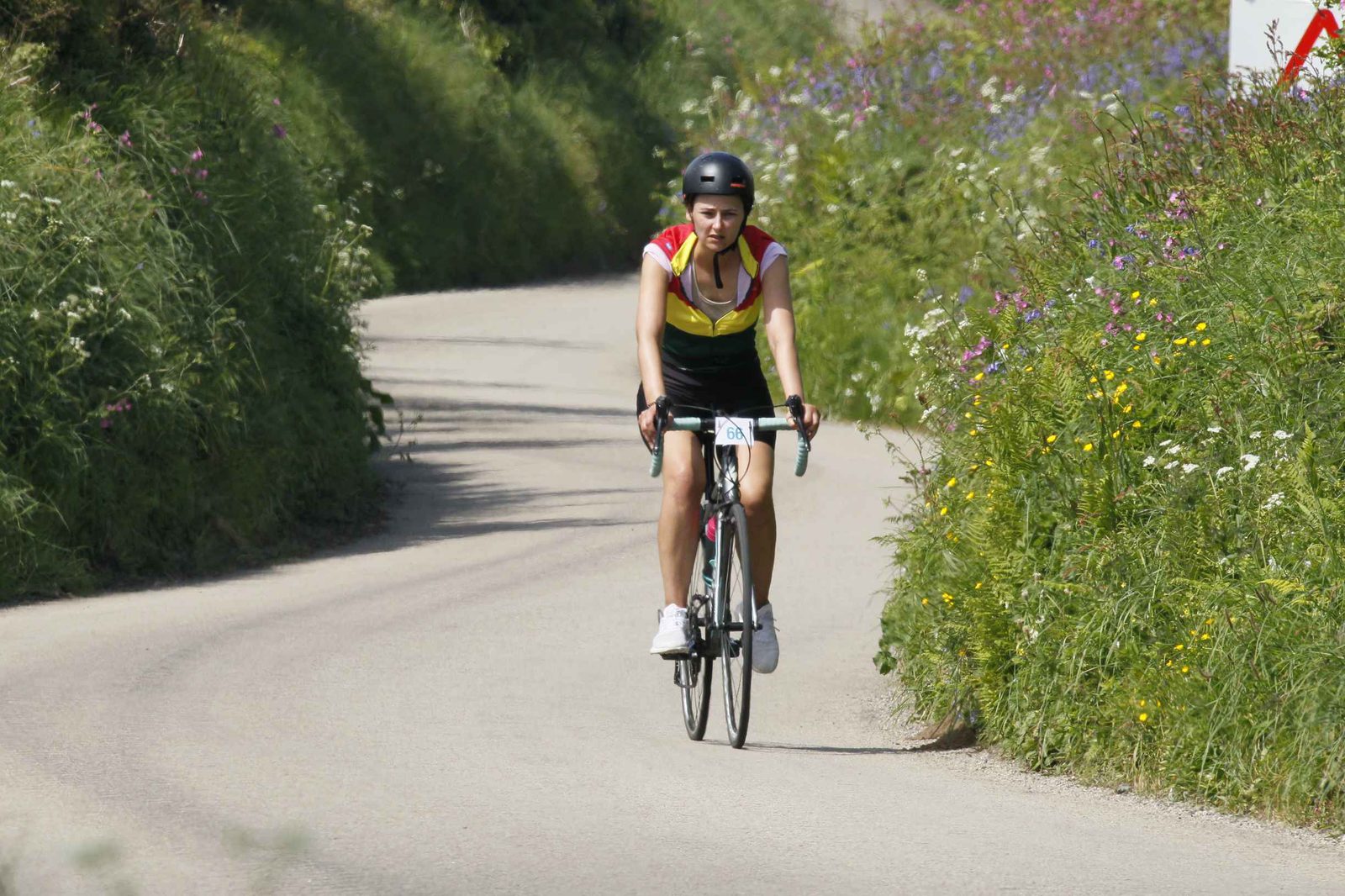 A person wearing a helmet and sports clothing is riding a road bike on a curvy, paved path flanked by vibrant greenery and wildflowers. The path ahead winds gently left amidst lush vegetation, suggesting a scenic cycling route.