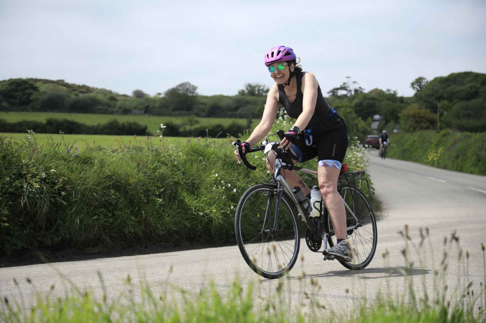 A person in a purple helmet and black cycling outfit is riding a road bike on a rural road surrounded by green fields and trees. Other cyclists can be seen in the distance on a sunny day.