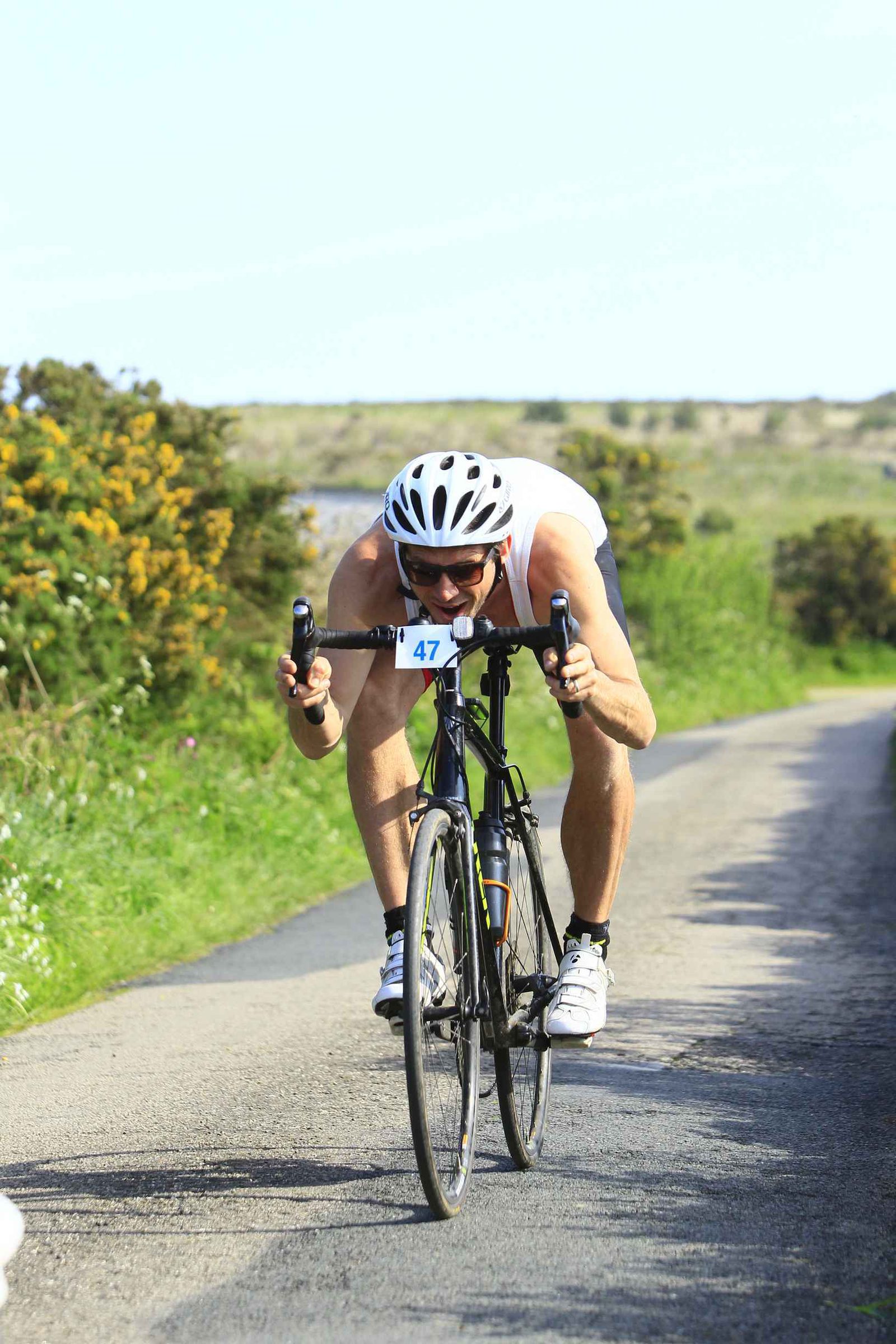 A cyclist wearing a white helmet, sleeveless shirt, and sunglasses rides a road bike at high speed on a narrow, sunny path surrounded by greenery and yellow flowers. The cyclist, focused with an aerodynamic posture, has a race number 47 attached to the front of the bike.