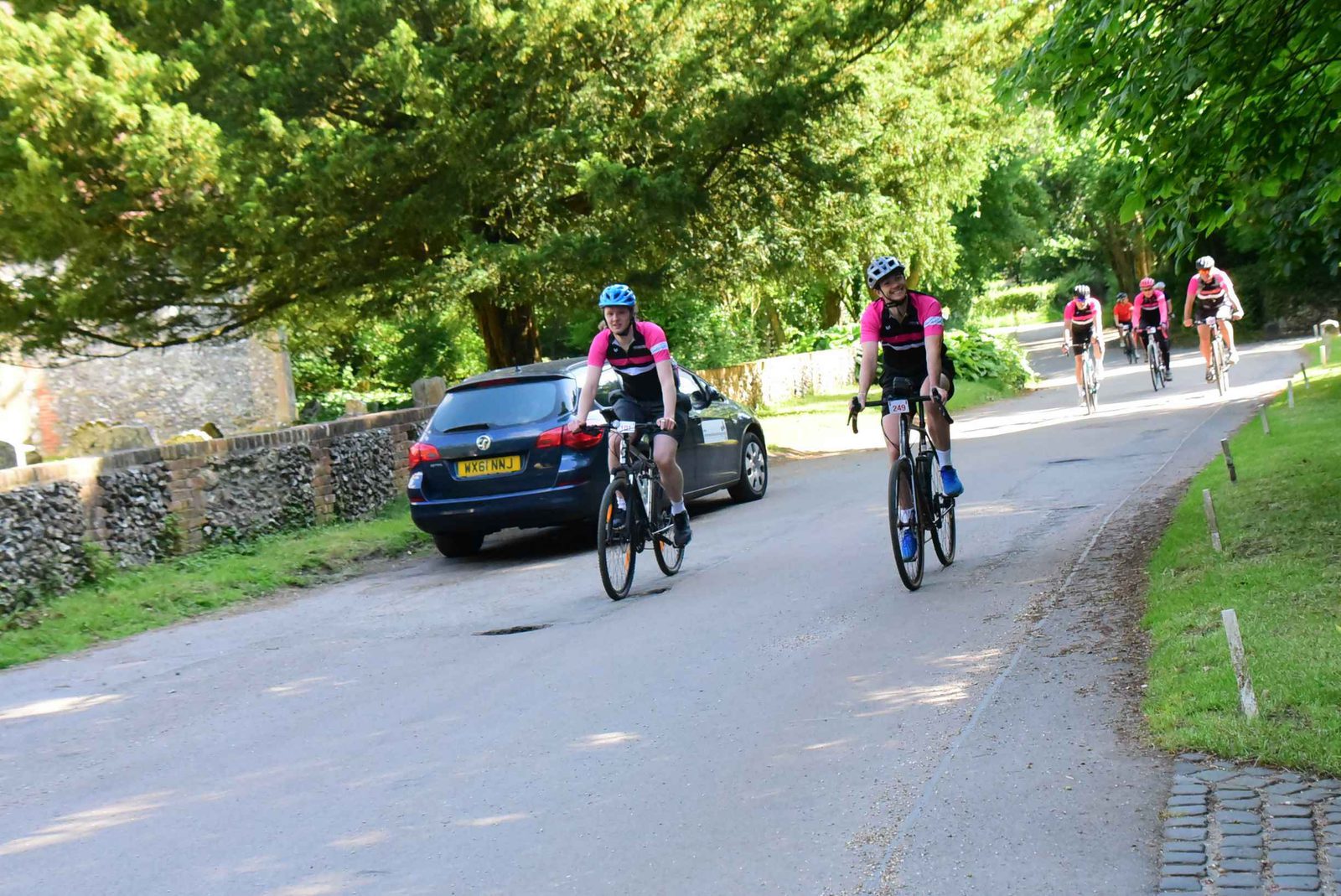 Five cyclists wearing pink and black outfits ride on a road through a green, wooded area. A parked car is visible on the left side of the road. Sunlight filters through the trees, casting shadows on the ground.