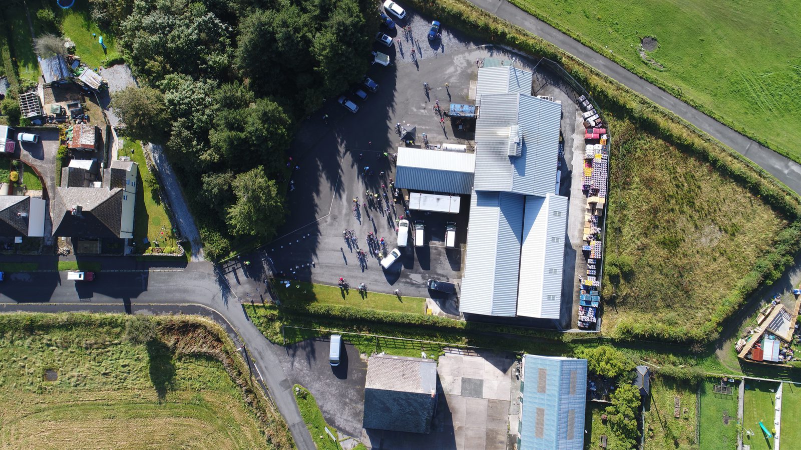 Aerial view of a large industrial building with a parking lot crowded with cars and people. Surrounding areas include residential houses, a green field, and roads. A smaller structure and a few scattered vehicles can also be seen nearby.