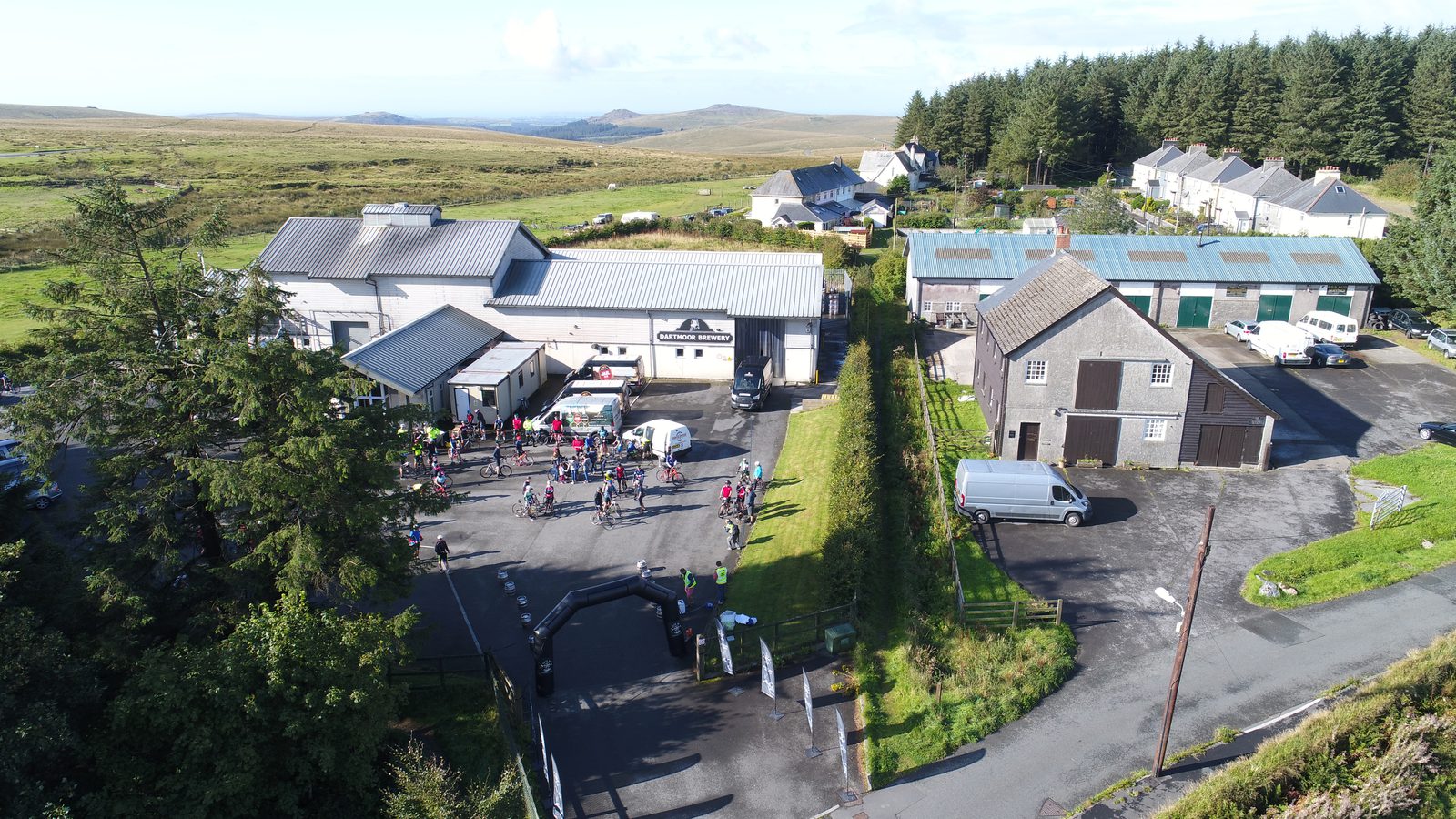 Aerial view of a rural event with people gathered in the courtyard of a farm-like property including several buildings, vehicles, and surrounding trees and hills. White tents and registration points are visible. Some people are running or walking on the surrounding road.