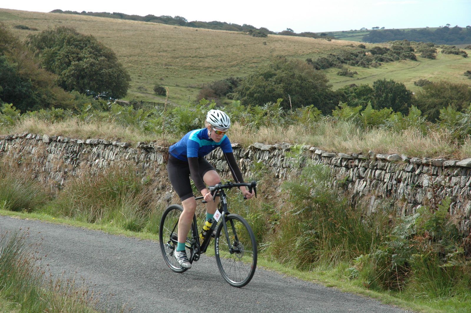 A cyclist wearing a blue and black outfit with a white helmet rides a bike on a paved road. In the background, there's a scenic landscape with grassy fields, bushes, and a stone wall. The sky is clear.