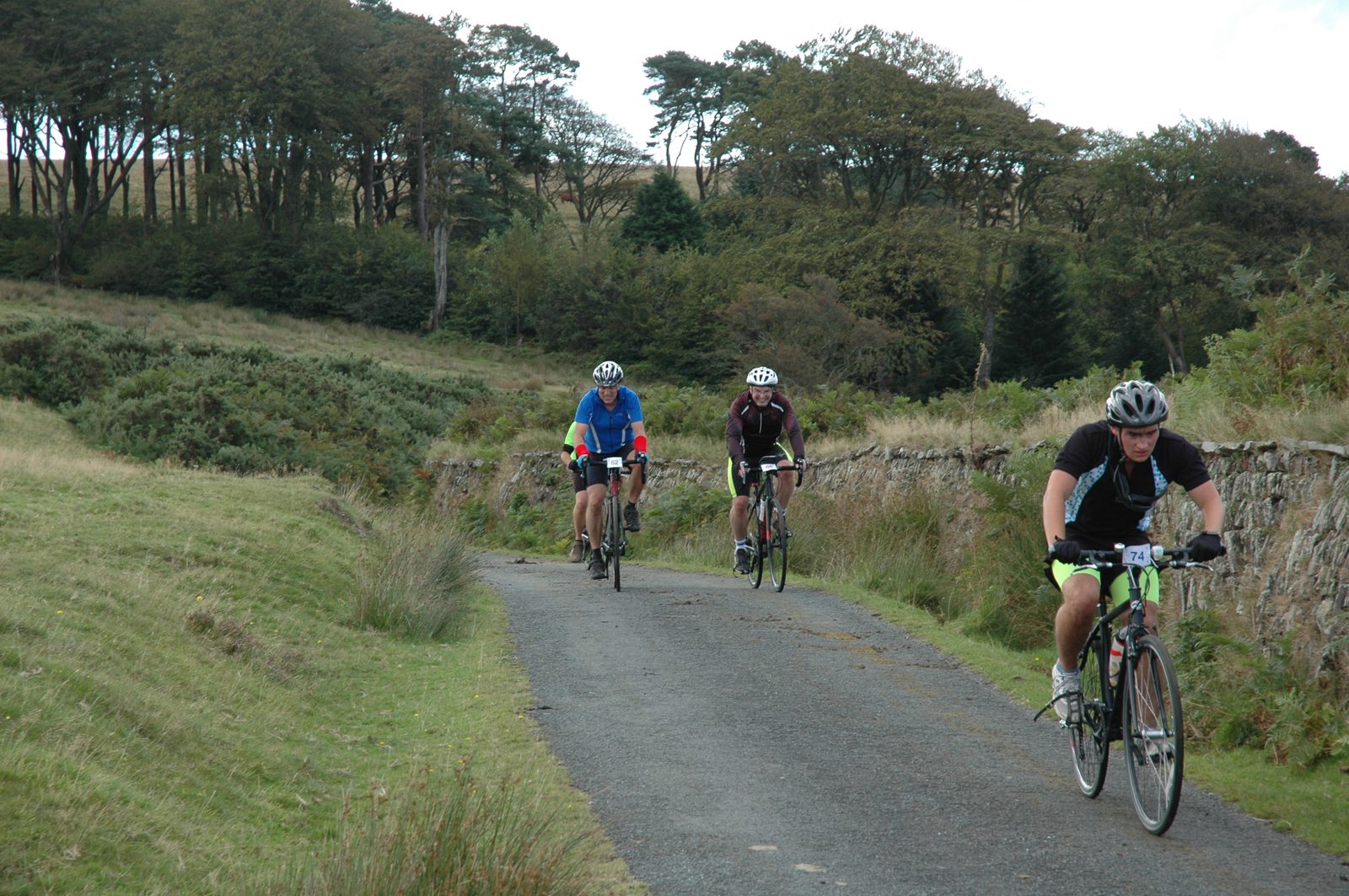 Three cyclists ride along a narrow country road flanked by grass and trees. The cyclists are dressed in different colored gear, with the leading rider wearing a black helmet and neon green shorts. The rural landscape features rolling hills and sparse woods.