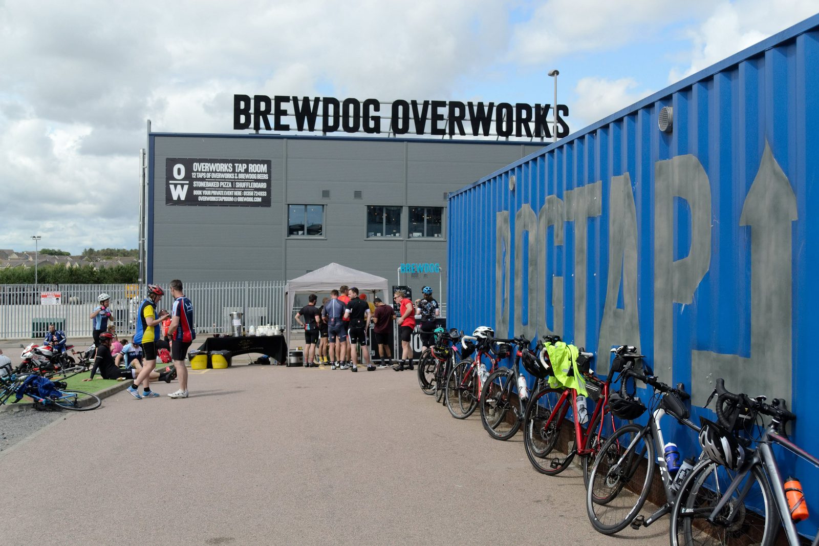 A group of cyclists gather outside BrewDog Overworks, a large grey building, near a blue shipping container with "DogTap" painted on it. Bicycles are lined up against the container. Some people are seated on benches, and a few others stand talking under a small canopy.