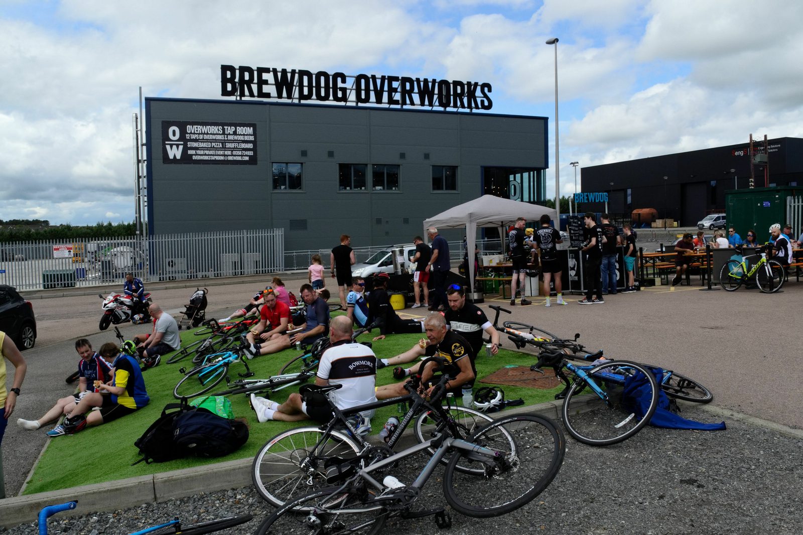 A group of cyclists is gathered outside the BrewDog Overworks building. Some are sitting on the ground, chatting, and resting, while others are standing near a white tent. Several bicycles are laid out on the ground nearby. The sky is partly cloudy.