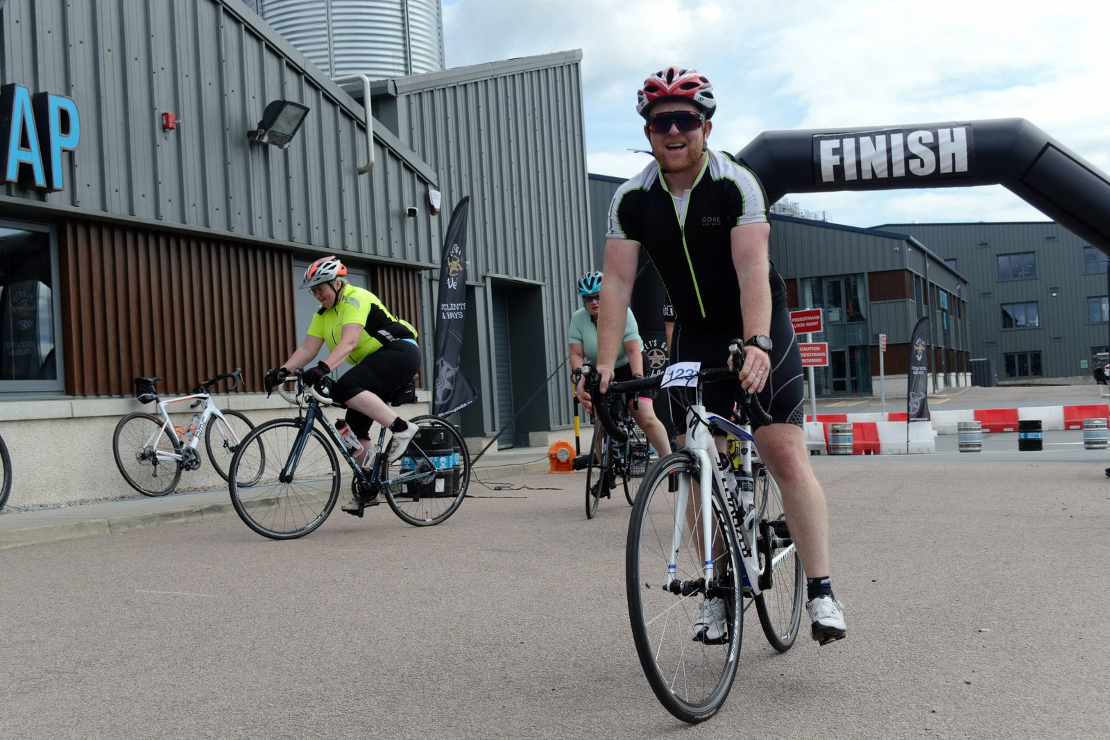 Cyclists at a race event's finish area. The person in the foreground is wearing a black cycling outfit and red helmet, crossing the finish line. Two other cyclists, one in a bright green top and another in a blue top, are seen in the background near the finish arch.