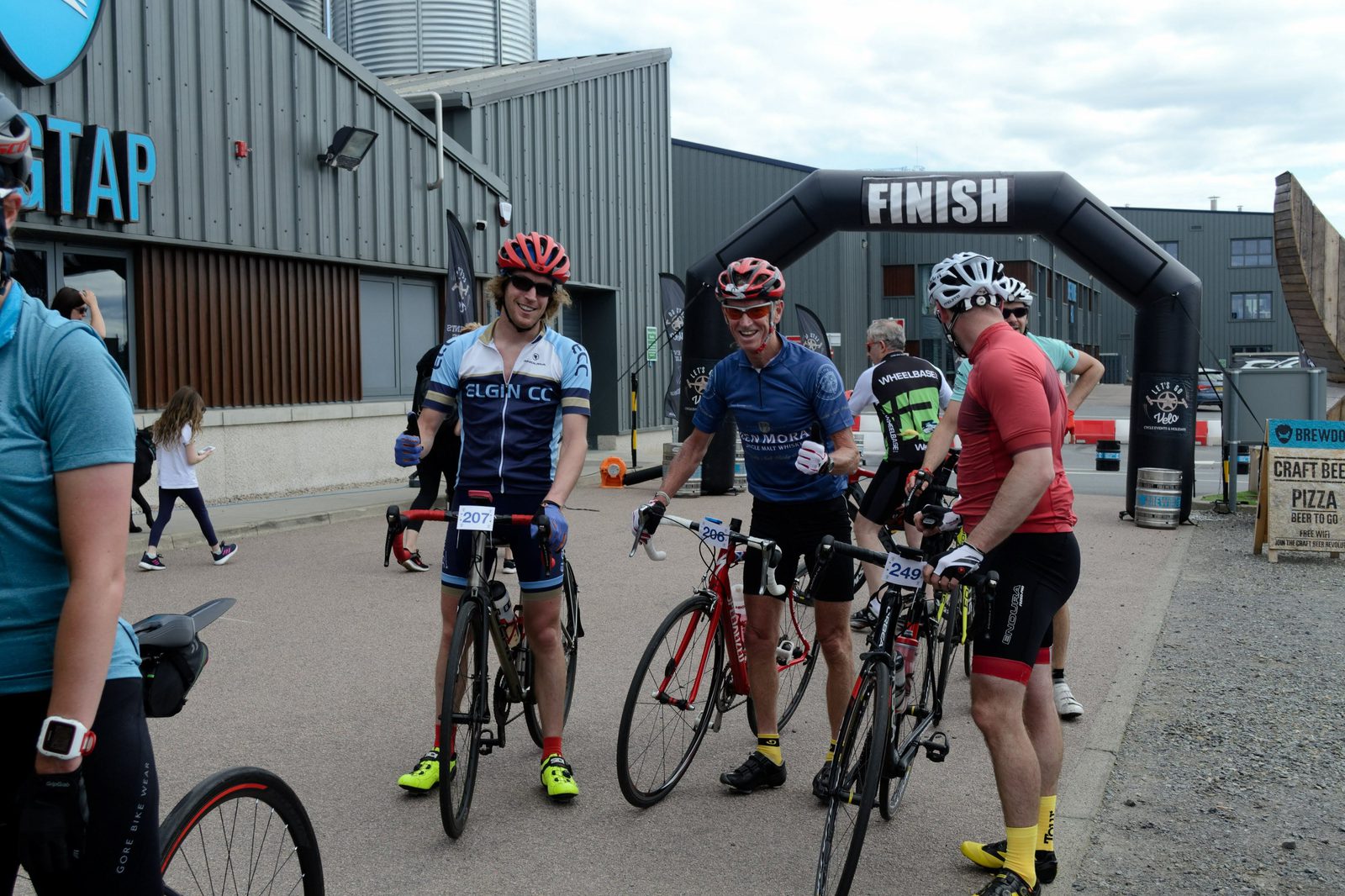 Cyclists gather near the finish line of a race, with three men in colorful cycling gear smiling and giving thumbs up, while holding their bikes. An inflatable "Finish" arch is in the background. Other participants and a gray building are also visible.