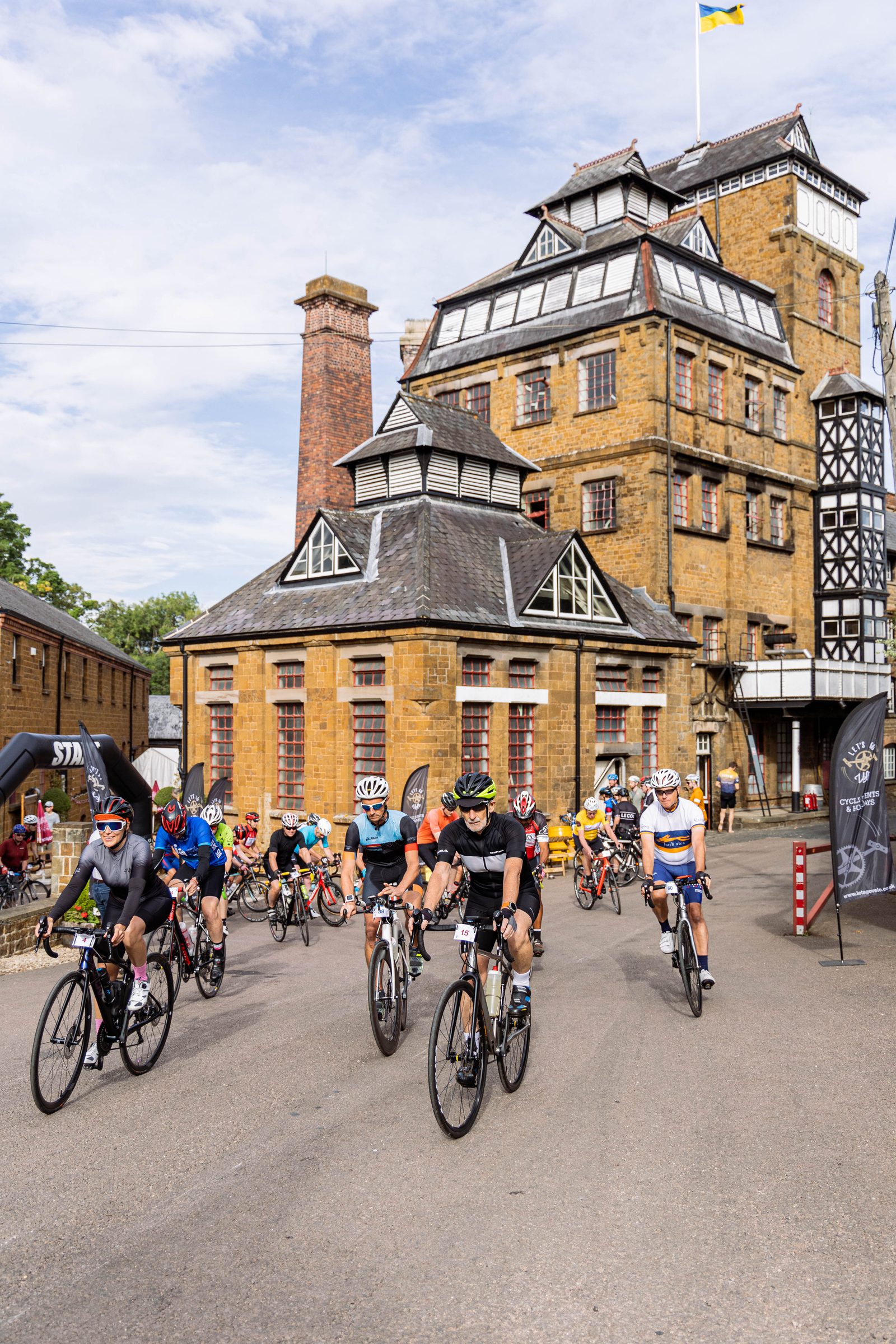 Cyclists ride past a historic, multi-story brick building with a large chimney, located outdoors on a sunny day. The building has a complex roof structure, and several cyclists are dressed in various bright athletic gear and helmets.