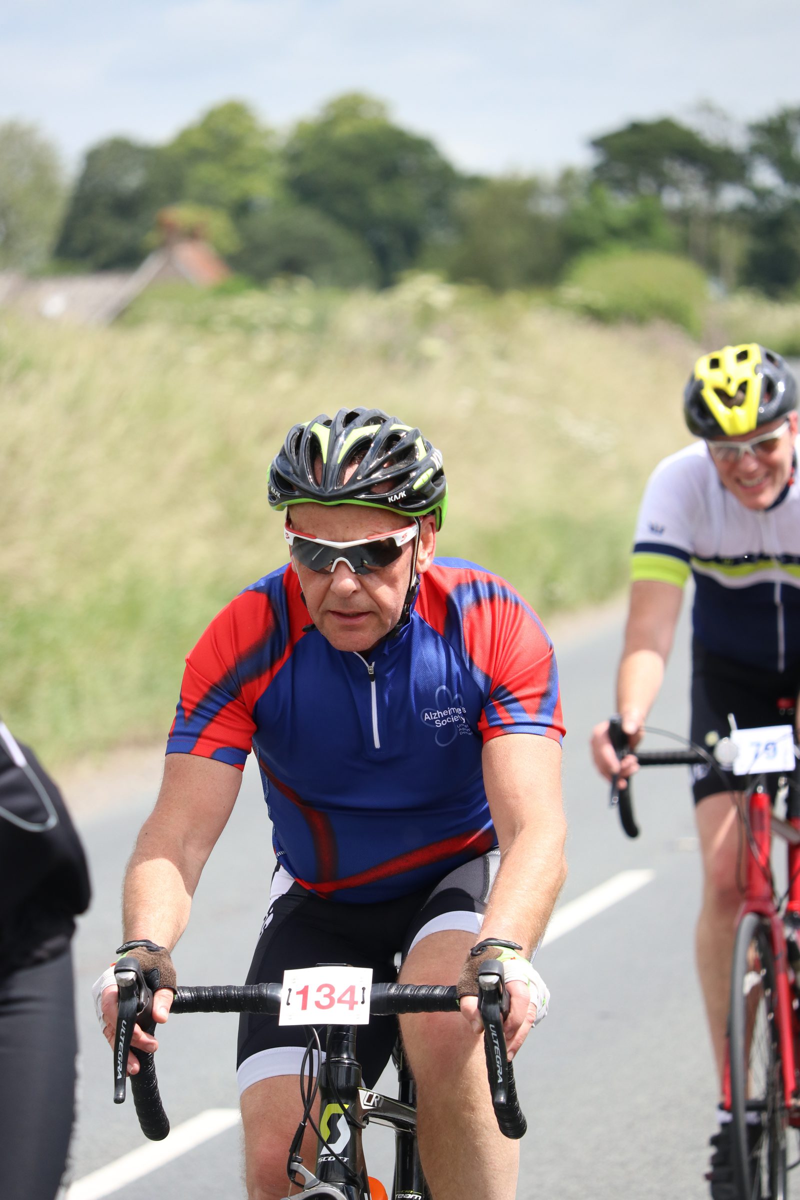 A cyclist wearing a red and blue jersey rides on a road, focused on the path ahead. He has sunglasses and a helmet on, with a race number 1341. Another cyclist in a white and blue jersey follows close behind, also wearing a helmet.