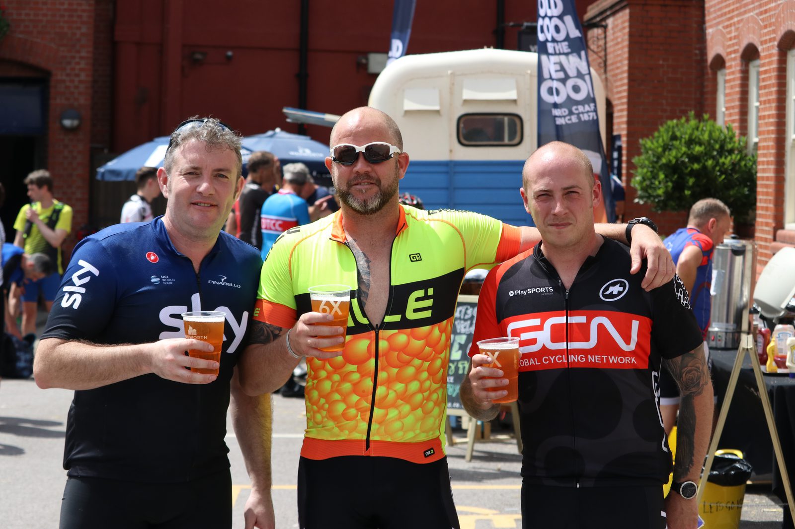 Three cyclists, dressed in colorful cycling jerseys, stand side by side holding pints of beer. They are outside near a building with other cyclists and people in the background. The man in the middle wears sunglasses, and all appear to be enjoying their time.