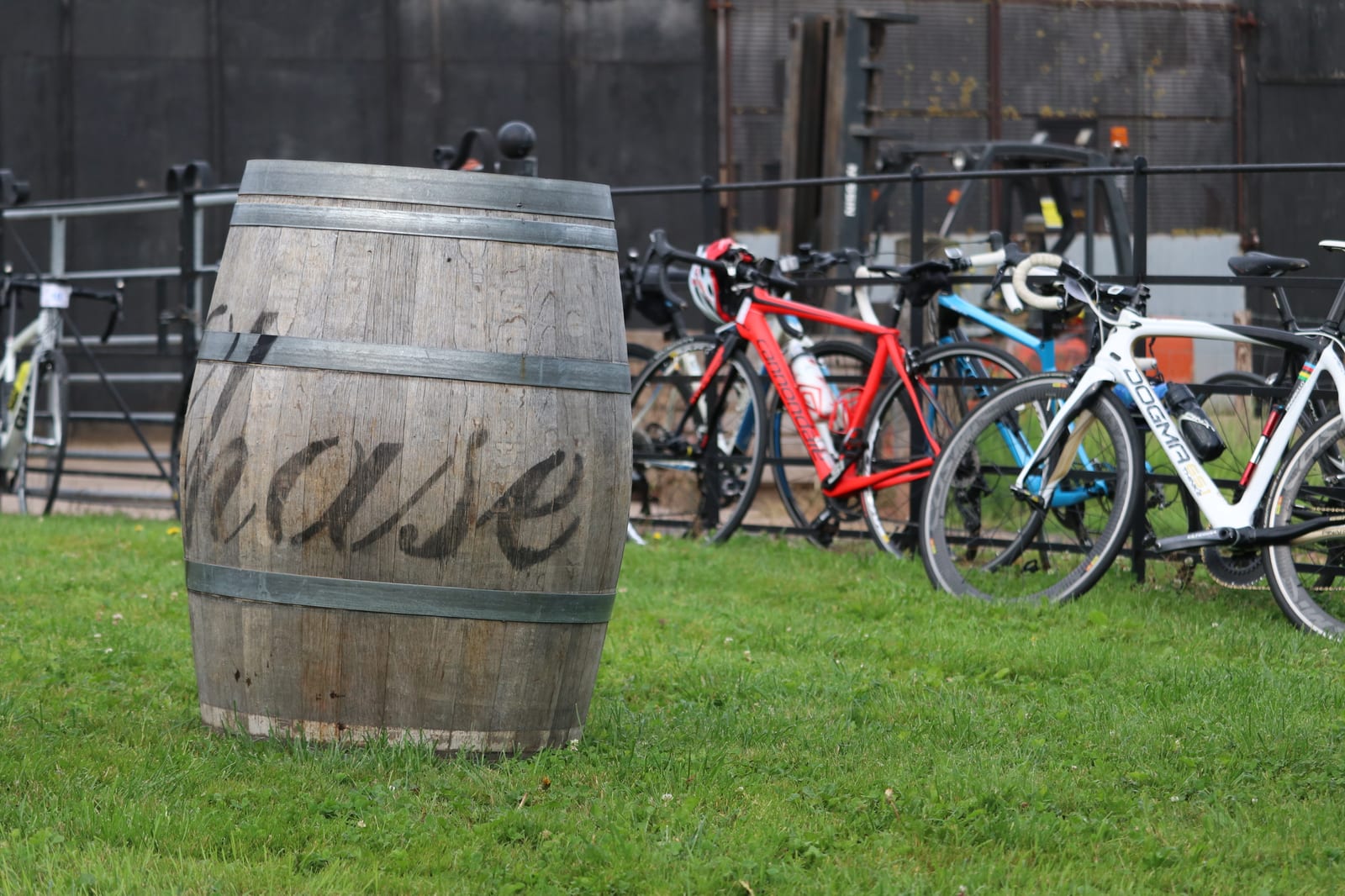 A wooden barrel with the word "chase" painted on it stands on a grassy area. In the background, several road bicycles are parked against a black metal fence. The scene appears to be outdoors, likely near a bike-friendly establishment.