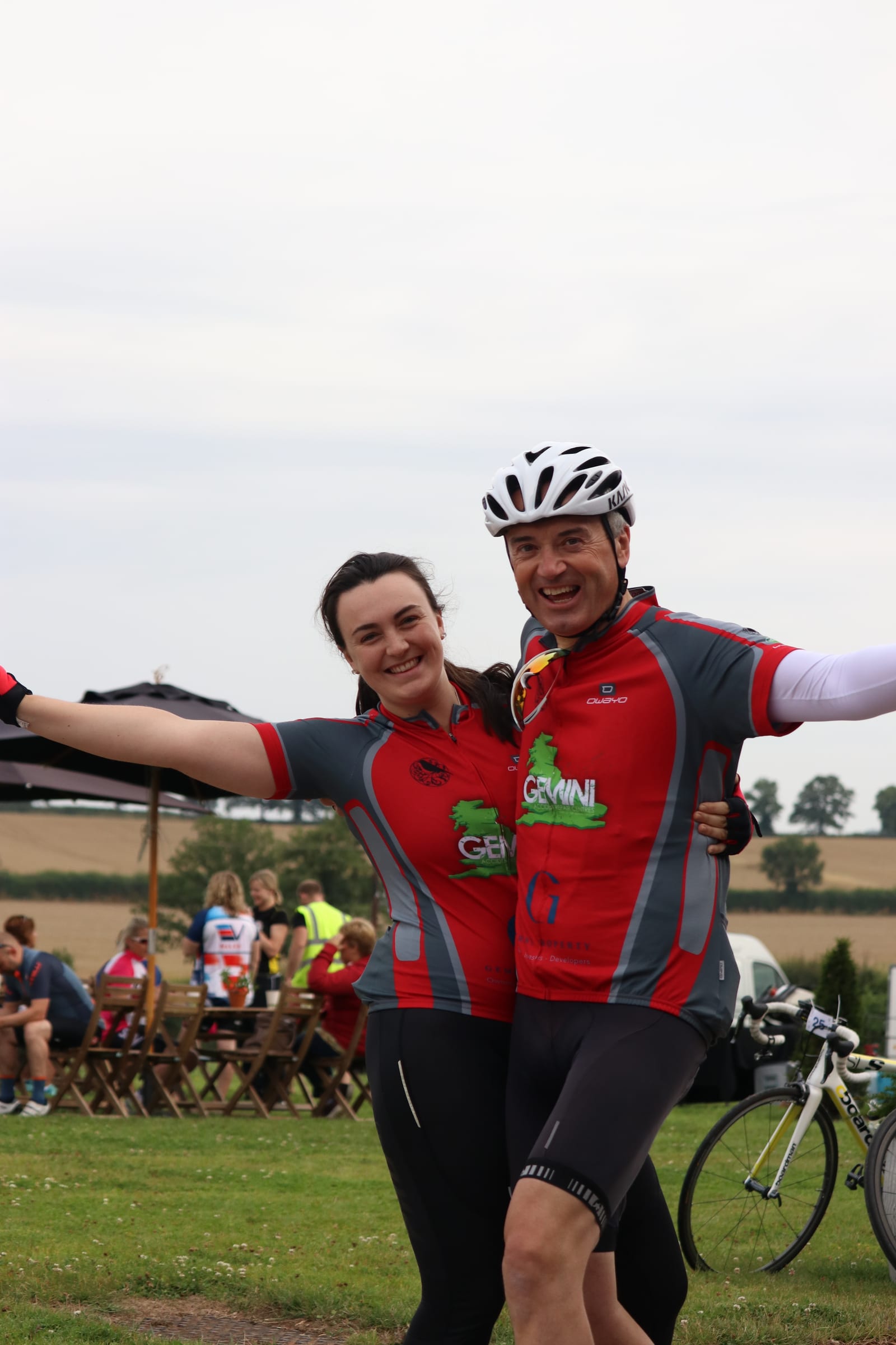 Two cyclists pose happily with their arms outstretched, wearing matching cycling jerseys. The background features other cyclists taking a break at picnic tables in a grassy area, with a rolling landscape and cloudy sky in the distance.