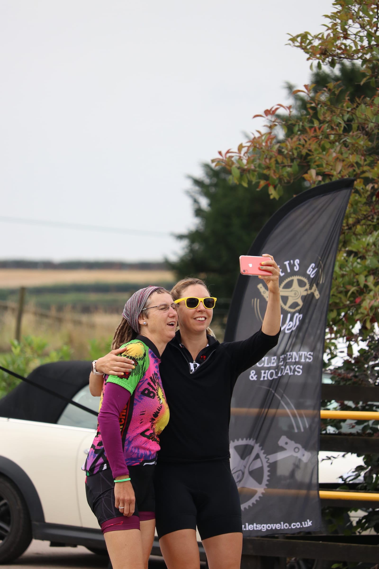 Two cyclists stand outdoors smiling and taking a selfie with a red smartphone. One wears a colorful jersey and headband, while the other wears black cycling gear and yellow sunglasses. Behind them is a black flag with the text "letsgovelo.co.uk" and some greenery.