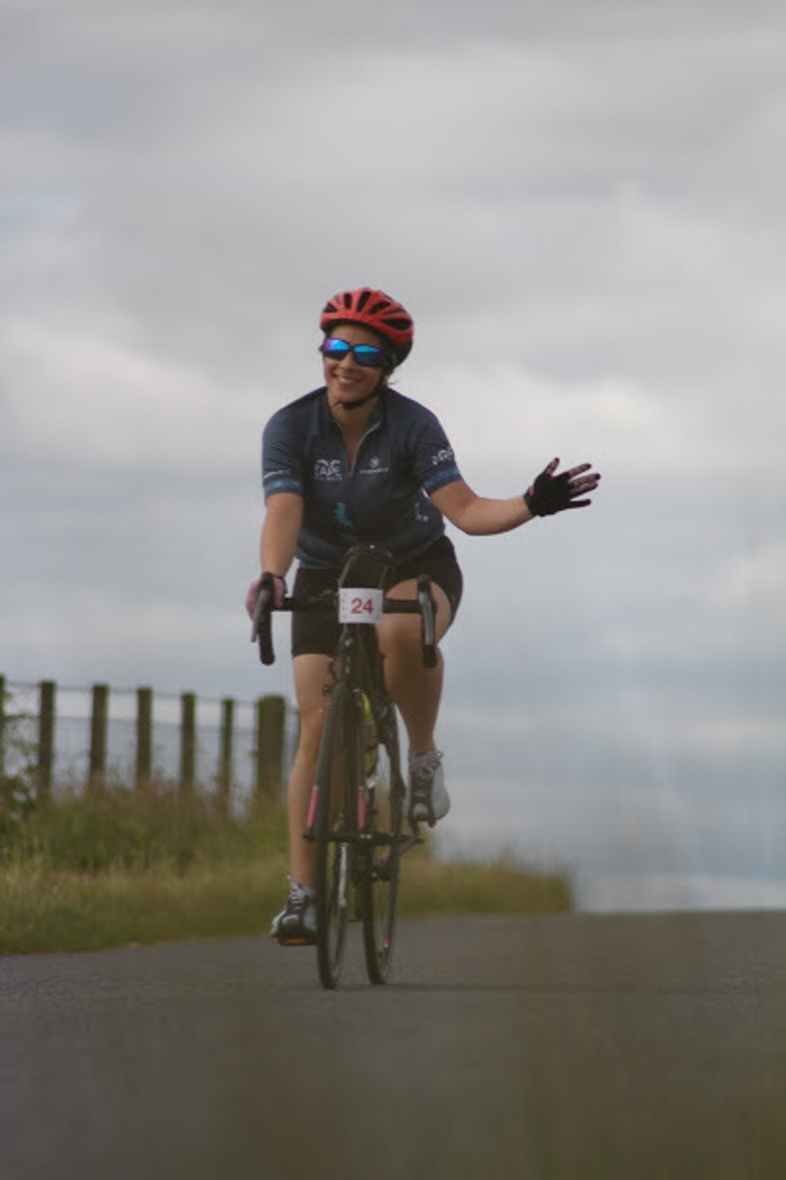 A cyclist wearing a red helmet, blue cycling jersey, and sunglasses rides along a road with one hand waving. The cyclist is smiling and has a race number 24 attached to the handlebars. The road is bordered by grass and a wooden fence.