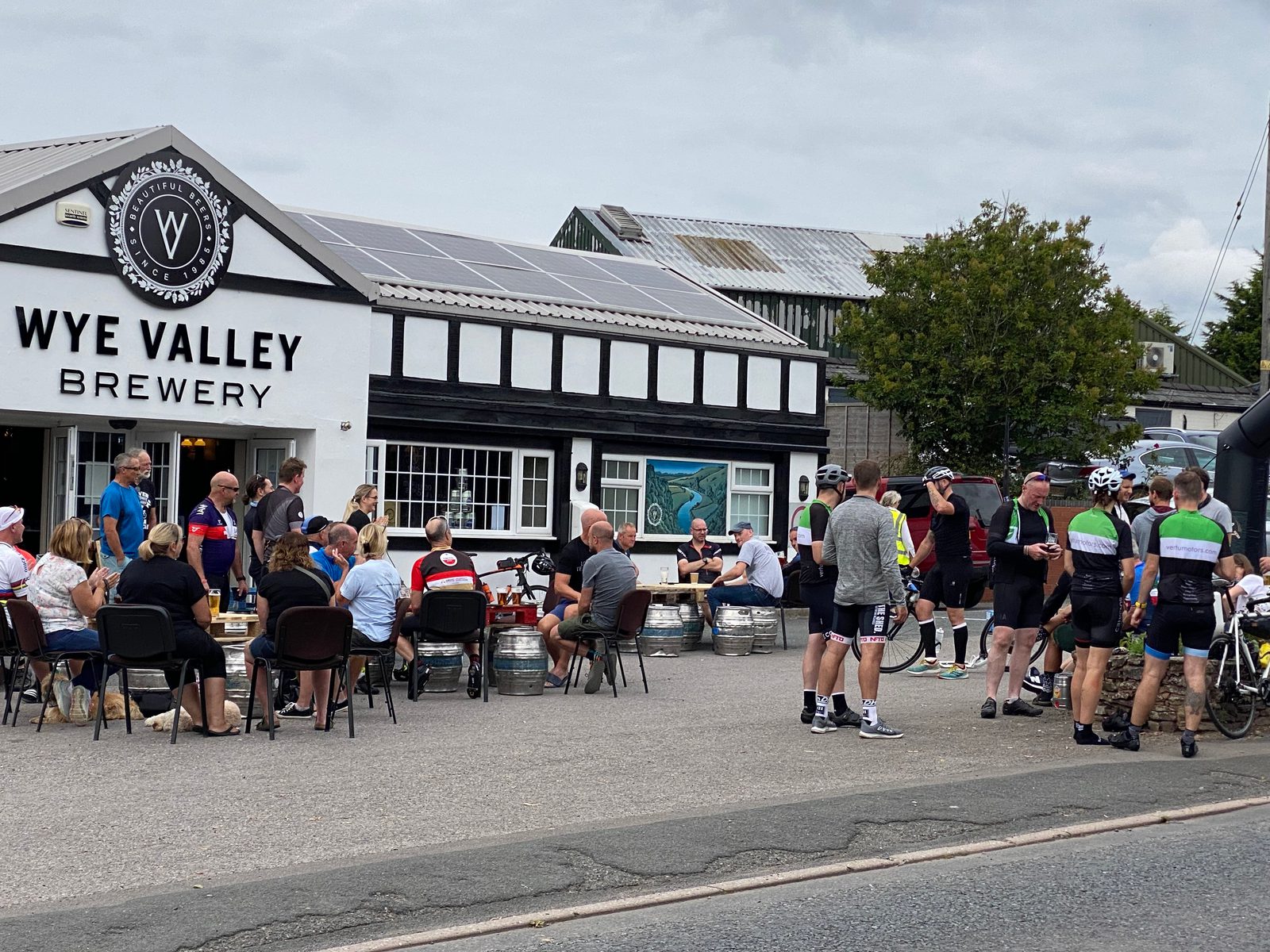 A group of people sitting and standing outside Wye Valley Brewery. Some are cyclists in gear, with bikes nearby. Others are seated at tables enjoying drinks. The building is white with black accents and has a sign with the brewery's logo. The sky is cloudy.