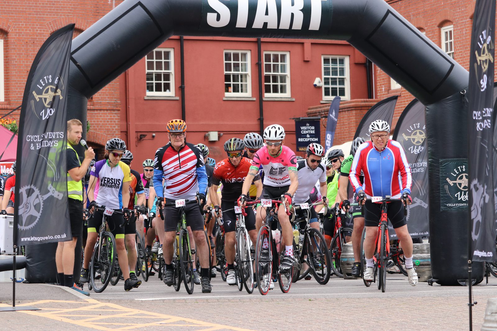 Cyclists lined up at a starting line under a black inflatable arch labeled "START." Riders wear various cycling gear and helmets, readying their bikes. Banners and a red brick building are in the background.