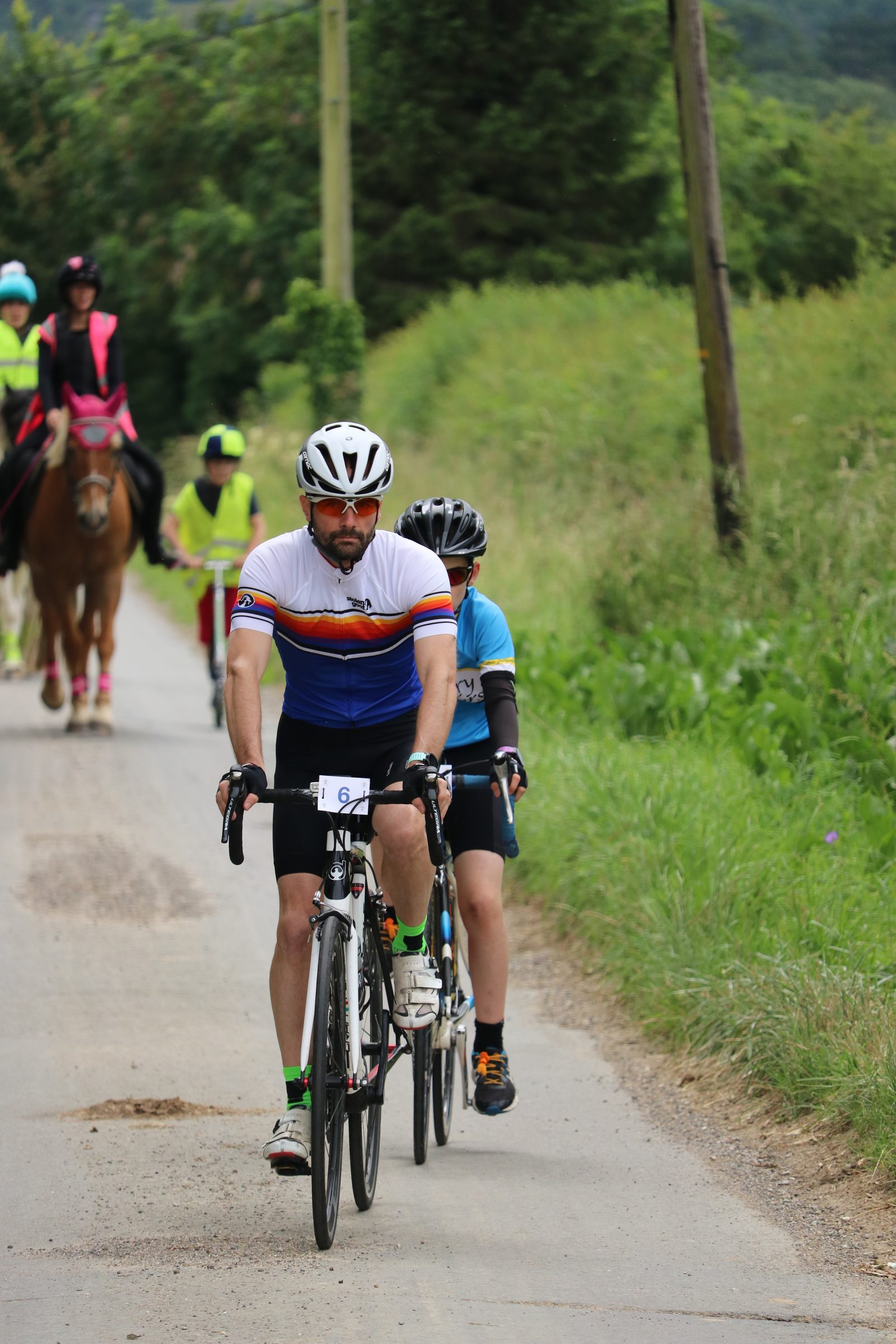 A man and a child riding road bicycles on a rural path lined with green foliage. The man wears a helmet and a white cycling jersey with blue, yellow, and red stripes, while the child follows closely in a blue and black jersey. Horse riders can be seen in the background.