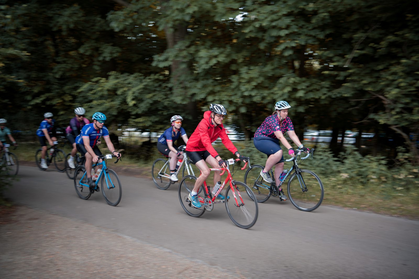 A group of cyclists ride on a paved path through a wooded area. They wear helmets and cycling attire. The cyclist in front is in a red jacket and white shorts. The others follow closely behind, dressed in various colorful outfits. Trees and greenery are visible in the background.