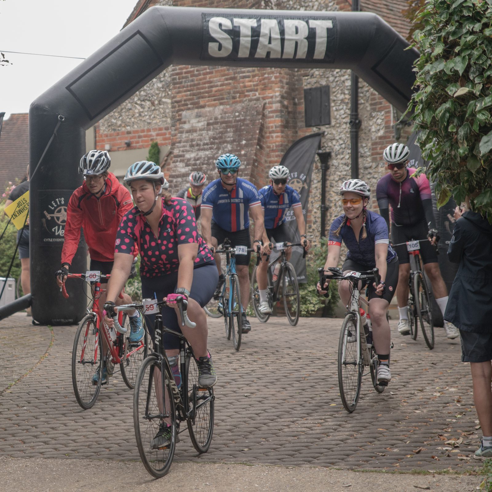 Cyclists in colorful gear begin a race under a large inflatable arch with "START" written on it. A stone building and lush greenery are in the background. The atmosphere is lively, with participants focused and ready to ride.