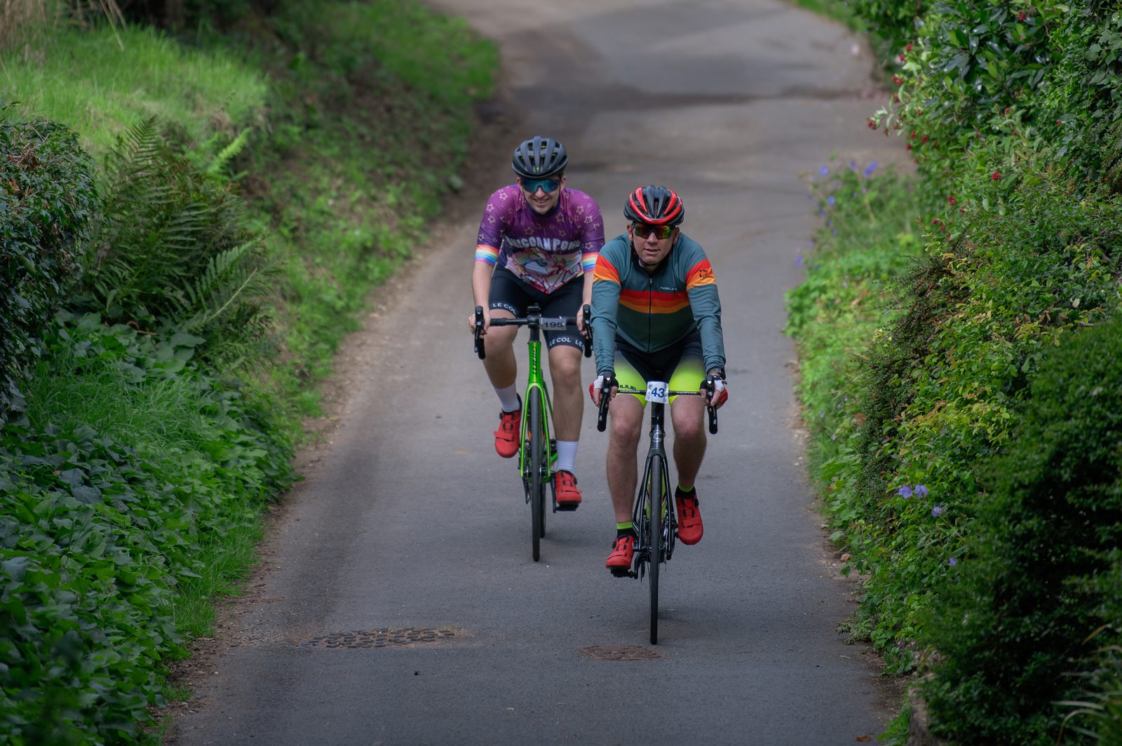 Two cyclists ride on a narrow, tree-lined road. The cyclist on the left is wearing a pink and purple jersey with green shoes, while the cyclist on the right is clad in a green and orange jersey. Both wear helmets and sunglasses as they pedal in a lush, green environment.