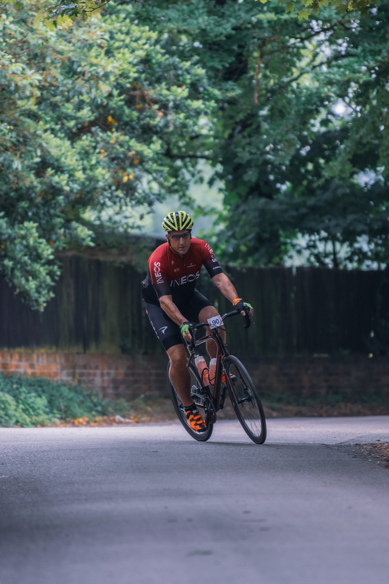 A cyclist wearing a red and black "Ineos" jersey and yellow helmet leans into a turn on a tree-lined road. The rider is on a sleek black road bike, and the background is lush with greenery, giving the scene a serene, natural setting.