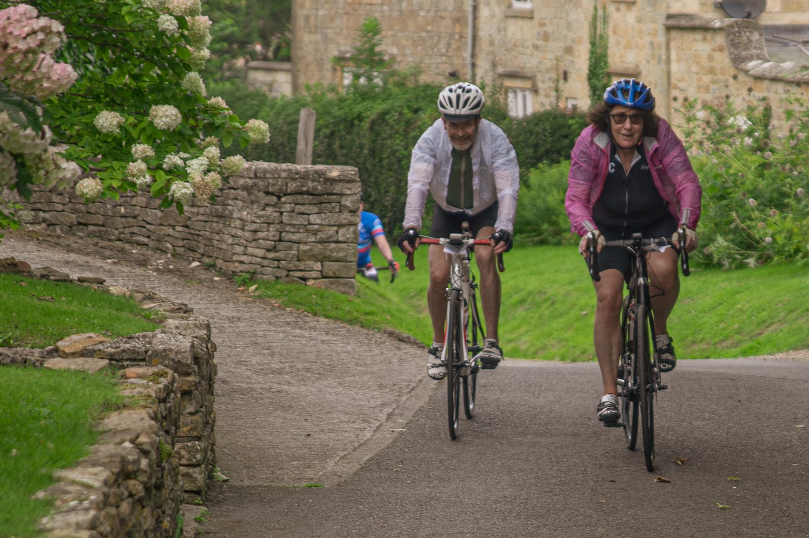 Two cyclists ride on a paved path through a quaint, countryside village. The first cyclist wears a white helmet and grey attire; the second wears a blue helmet and pink jacket. Both smile as they cycle past stone walls and green foliage, with a stone house in the background.