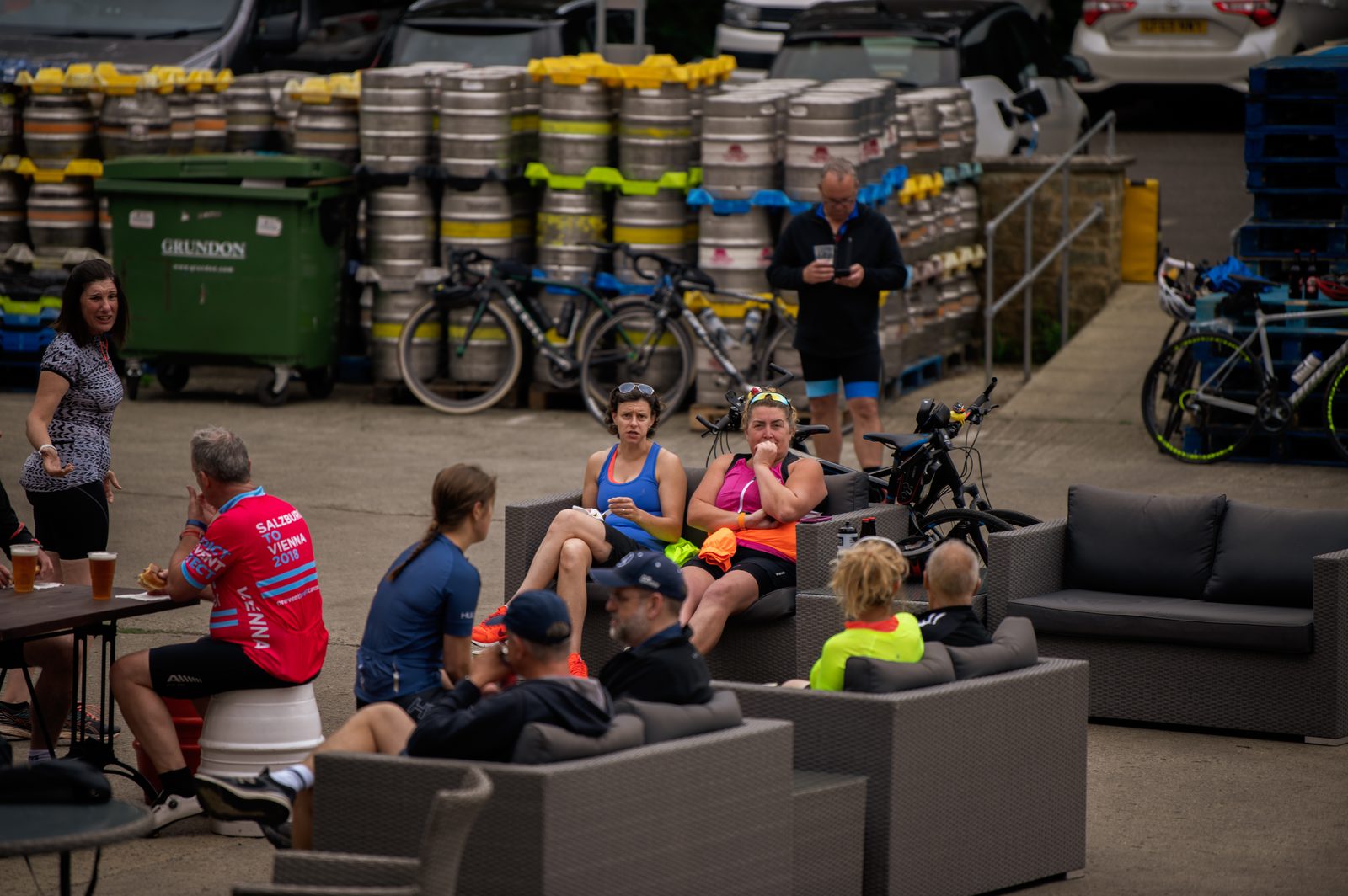 A group of cyclists in colorful gear relax on sofas and chairs outdoors near stacked beer kegs and parked bicycles. They are engaged in conversation, with some holding drinks. The background includes recycling bins and more bikes, suggesting a casual social gathering post-ride.