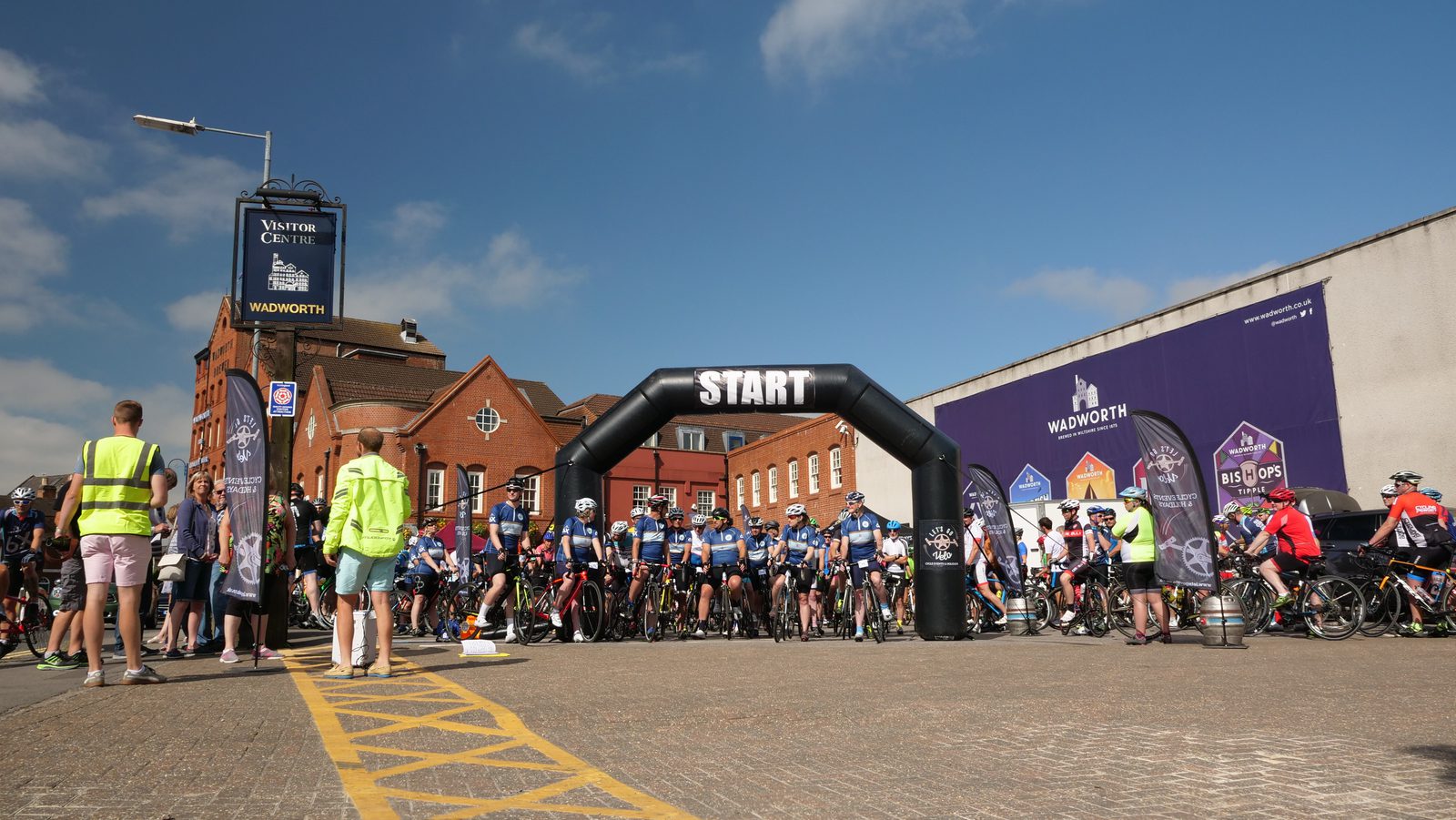 A large group of cyclists is lined up at the starting line under an inflatable arch marked "START" on a sunny day. The event takes place in an urban area with red brick buildings and banners for "Wadworth." People in high-visibility vests are overseeing the scene.