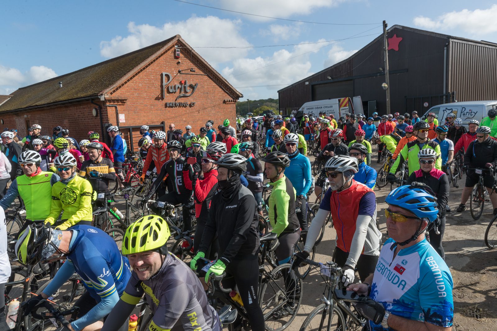 A large group of cyclists in colorful gear gather outside Purity Brewing Co. on a sunny day. They are preparing for a ride, with some chatting and others adjusting their equipment. The sky is clear with light clouds, and buildings are visible in the background.