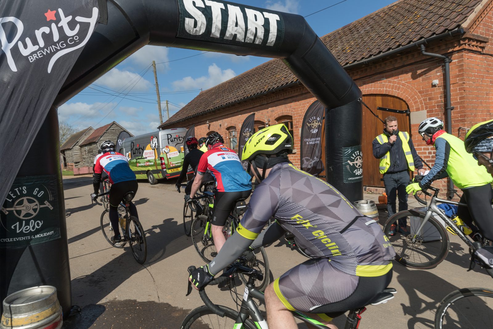 Cyclists in various colorful jerseys begin a bike race under an inflatable "START" archway near brick buildings. A Purity Brewing Co. van and several people, including a man in a yellow vest, are in the background on a sunny day.