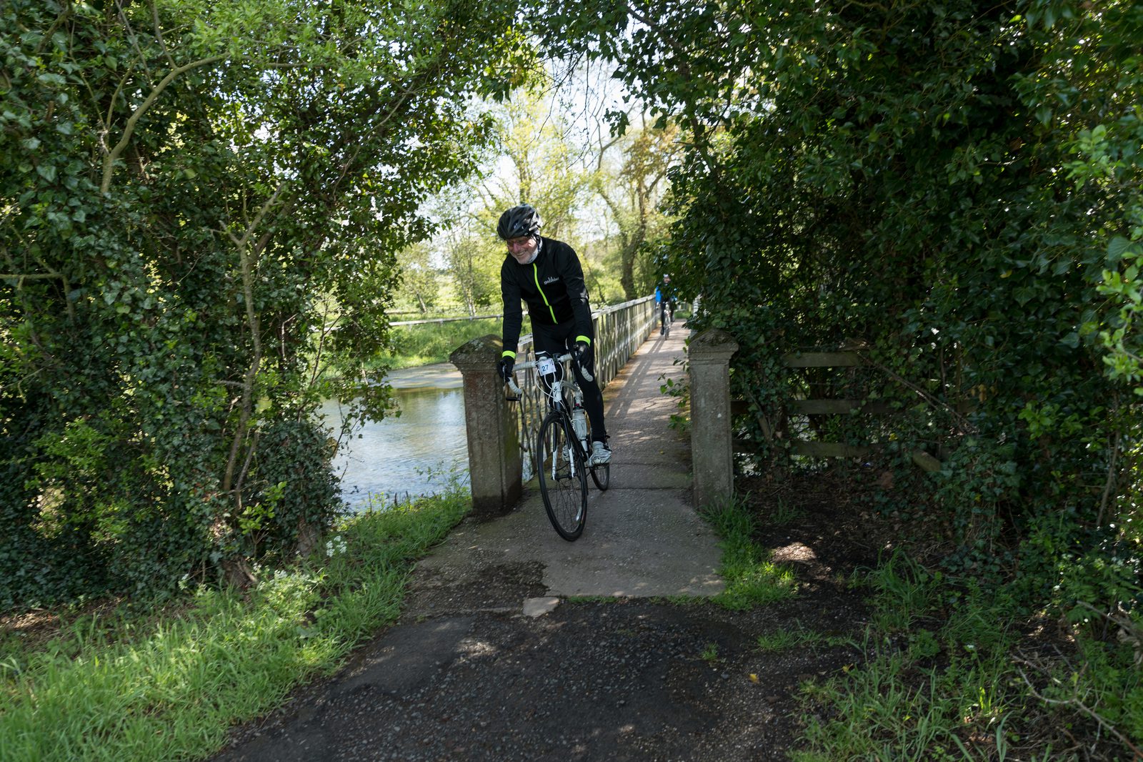 A cyclist in black and neon yellow gear rides a road bike on a narrow, shaded path surrounded by lush green foliage. A river and bridge are visible in the background, suggesting a serene outdoor setting. The rider appears focused, enjoying the scenic route.