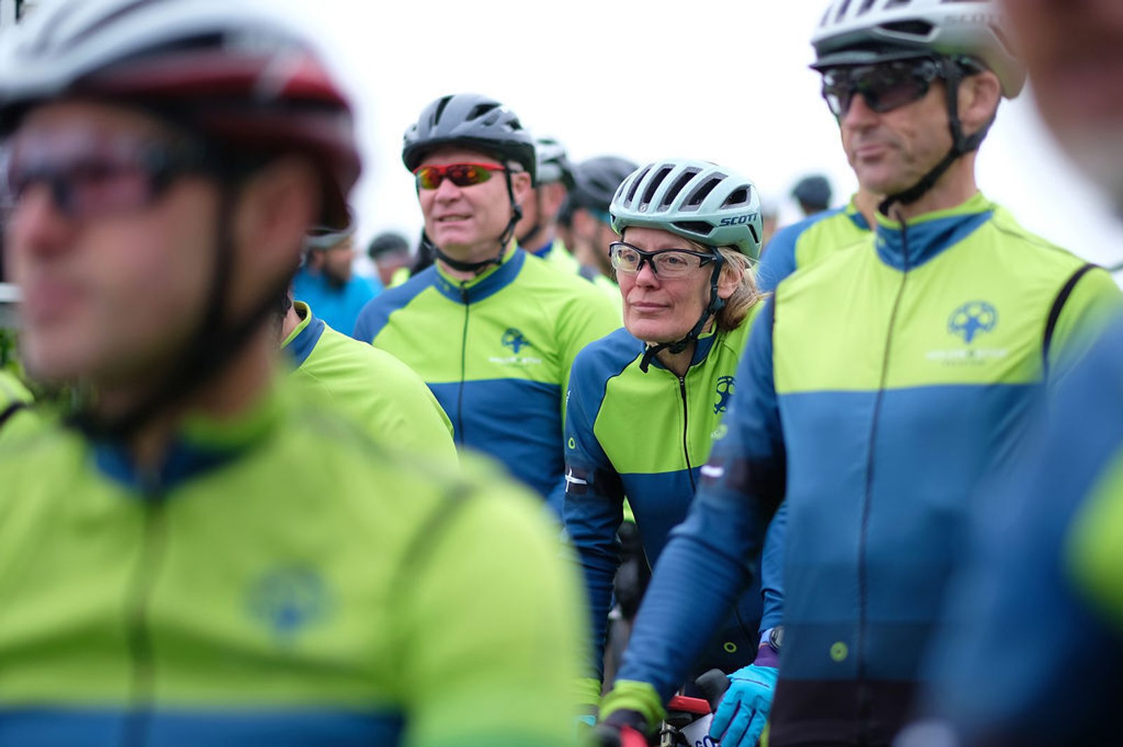 A group of cyclists wearing matching green and blue jerseys and helmets are gathered closely together. One cyclist, a woman with glasses, is in focus while the others are slightly blurred in the foreground and background. They appear to be participating in an event.