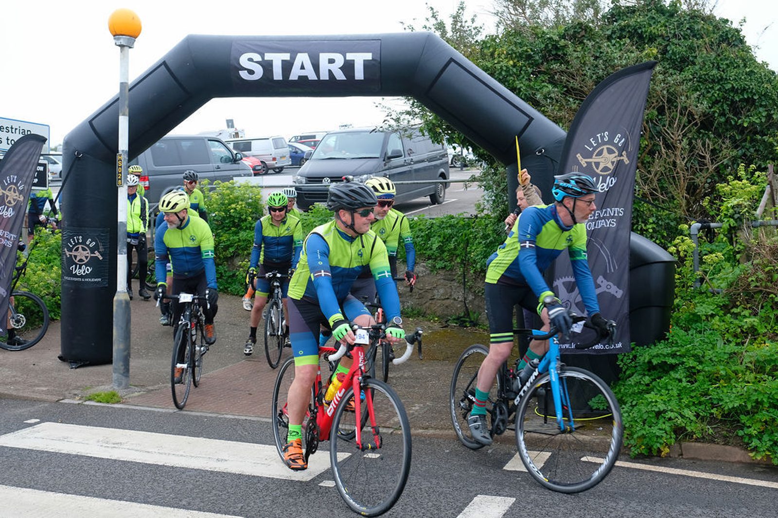 Cyclists in green and blue jerseys begin a ride, passing under a black inflatable arch with "START" written on it. They are on a road by a parking area, with several more cyclists behind them. Black banners flank the arch, and a crossing is visible in the foreground.