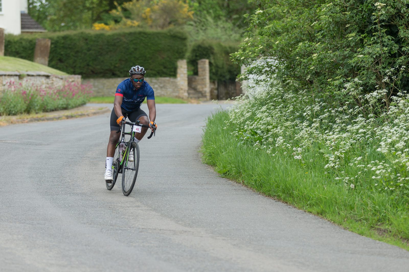 A cyclist wearing a blue jersey, black shorts, and white socks rides along a curved, scenic rural road bordered by lush greenery and flowering plants. The cyclist appears focused, with a helmet and sunglasses on. In the background, there are stone walls and trees.