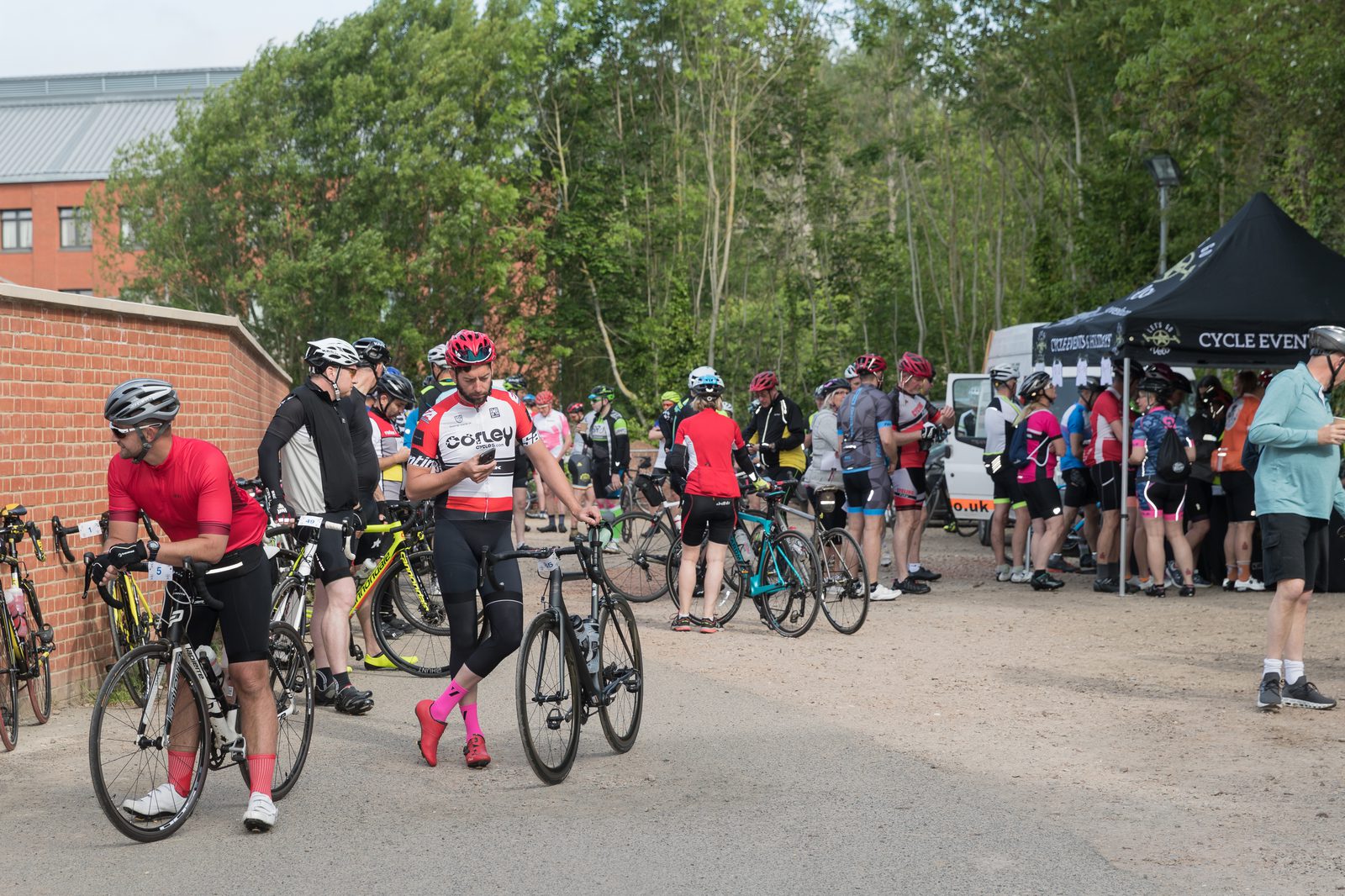 Cyclists gather at a rest stop during an event. Several people stand around with their bikes, chatting and checking phones. A tent labeled "Cycle Event" and a refreshments truck are in the background among trees and a brick wall. The area is busy and lively.