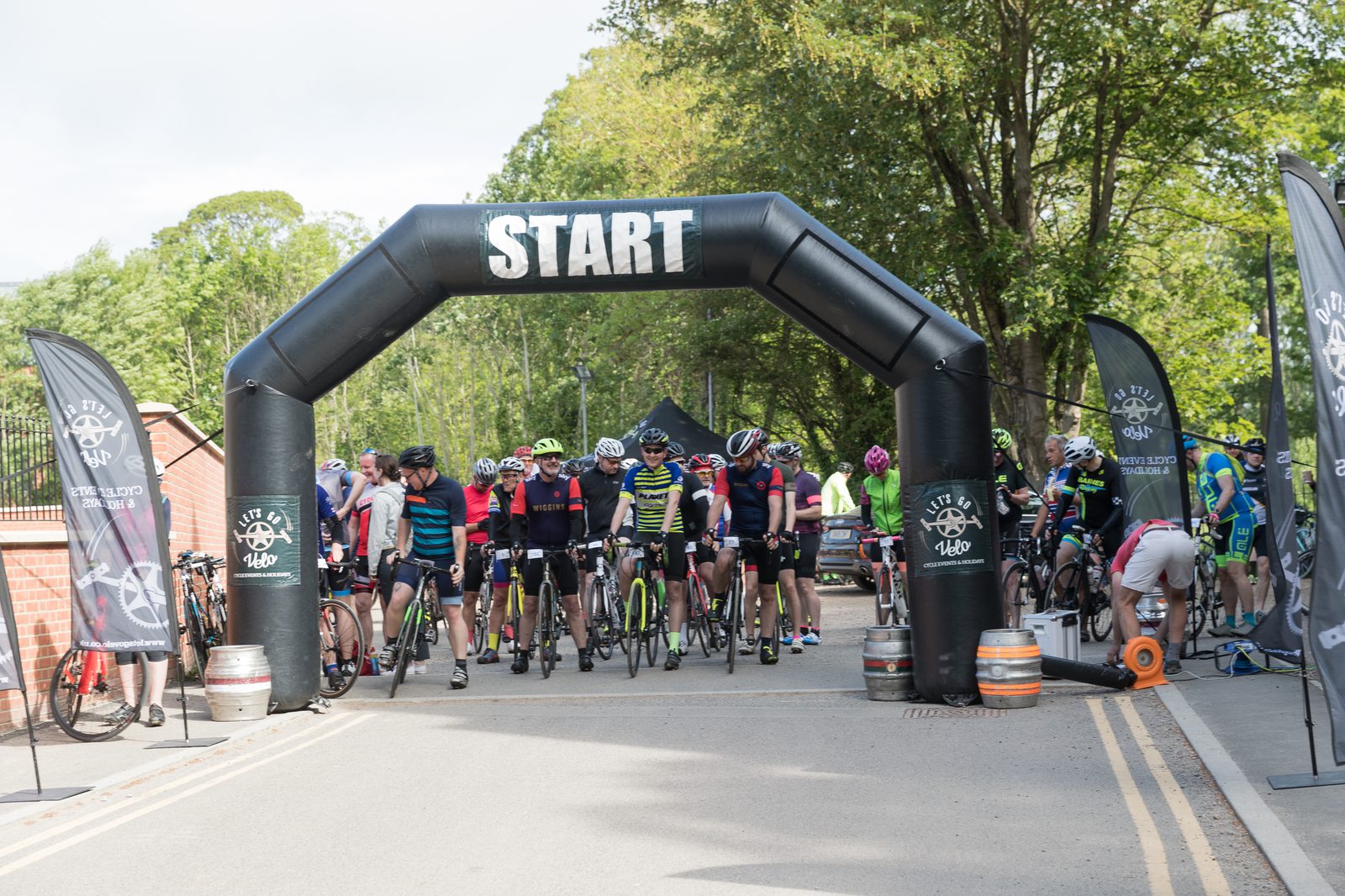 Cyclists gather at a starting line under an arch labeled "START" for a race. They are in colorful cycling gear and helmets, ready to begin. Trees and greenery are in the background, and flags with logos flank the starting area. A photographer is capturing the scene.